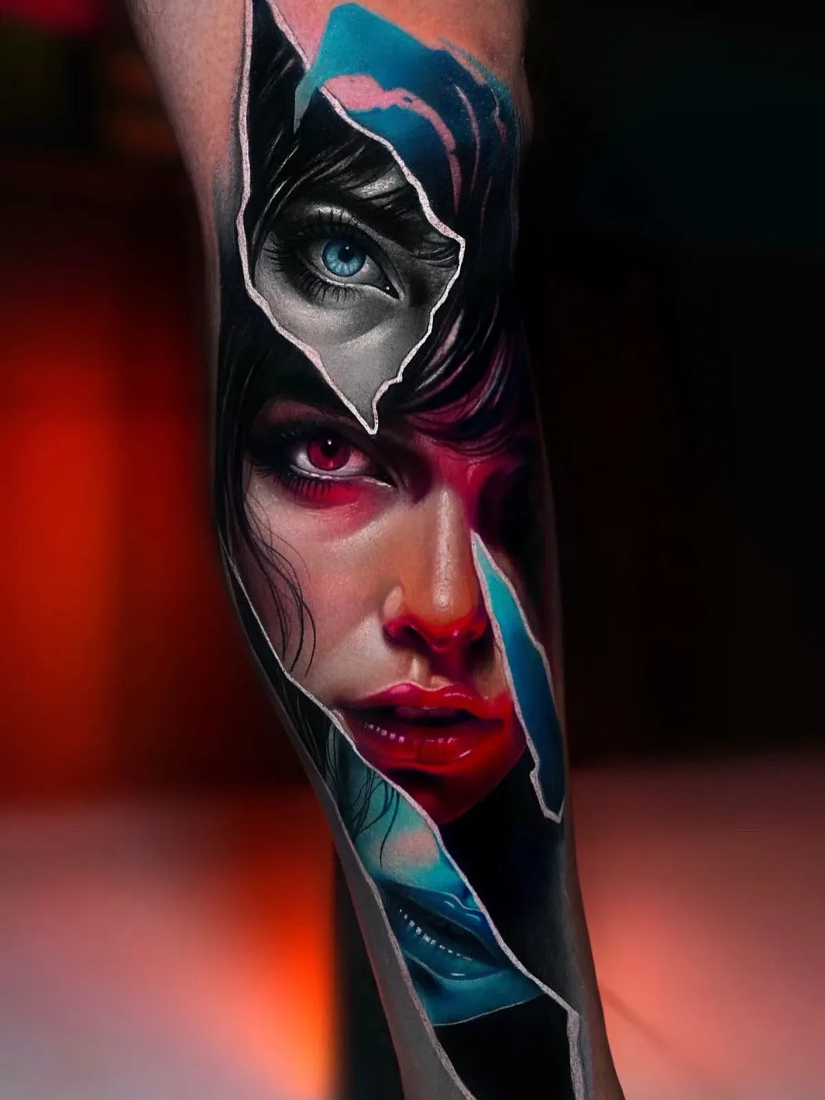 A hyperrealistic tattoo on a leg depicting fragments of faces with a torn-skin illusion; a blue eye sits in the upper section, a red eye and glossy red lips dominate the centre, and blue lips are glimpsed through jagged tears in the lower layer.