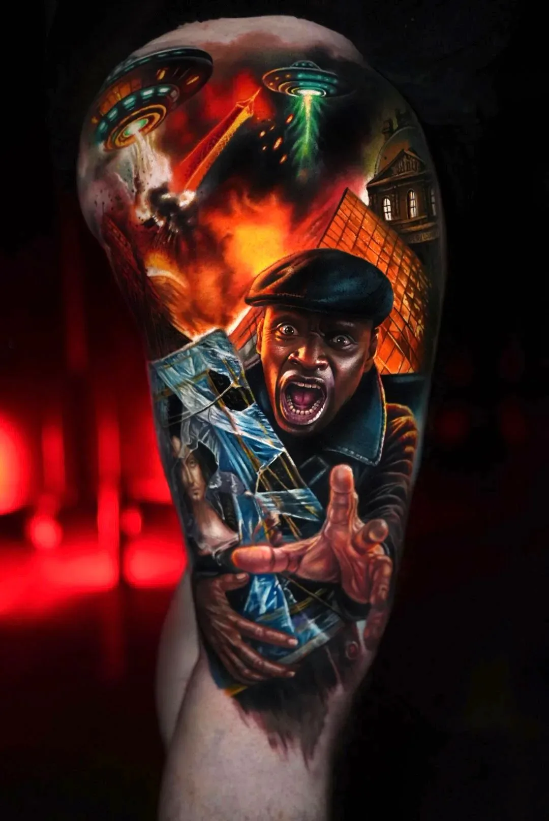 A hyperrealistic colour tattoo showing a Black man's face contorted in terror, reaching outward while holding a broken picture frame containing a woman's portrait; two UFOs emit coloured beams above a burning city with a pyramid and classical building in the background.