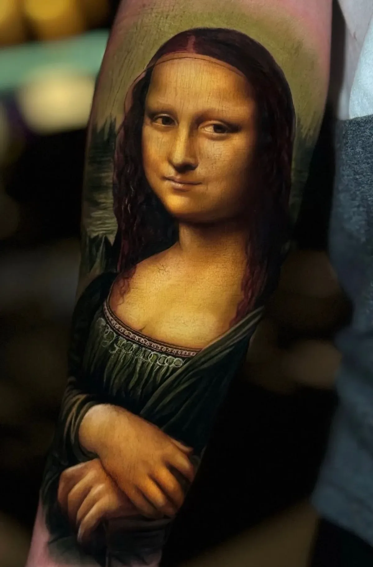 A hyperrealistic tattoo of the Mona Lisa on an arm, depicting the subject's face, clasped hands and misty landscape background in warm earthy tones with fine cracked-paint texture.