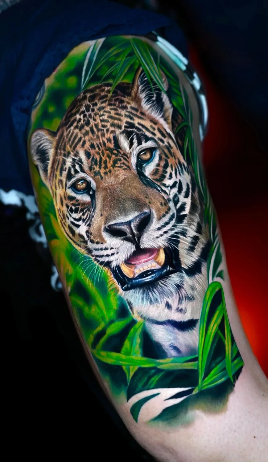 A hyperrealistic colour tattoo of a jaguar's head surrounded by tropical leaves; the jaguar faces forward with its mouth slightly open, amber eyes catching the light against a dark background.