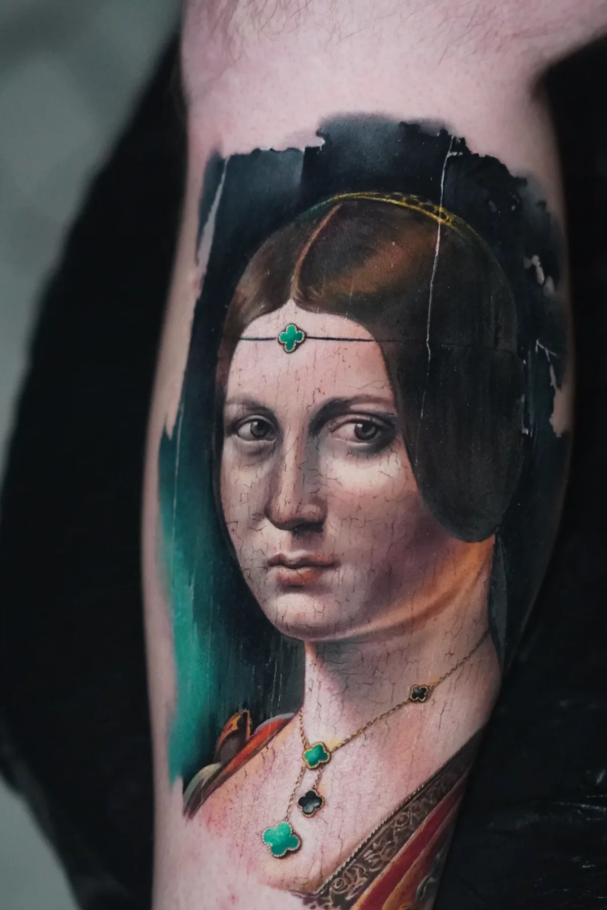 A hyperrealistic tattoo on an arm depicting a young woman in Renaissance portrait style with dark hair, a gold headband and a necklace bearing green and black clover pendants; fine cracked-paint texture overlays the skin tones against a dark teal background.