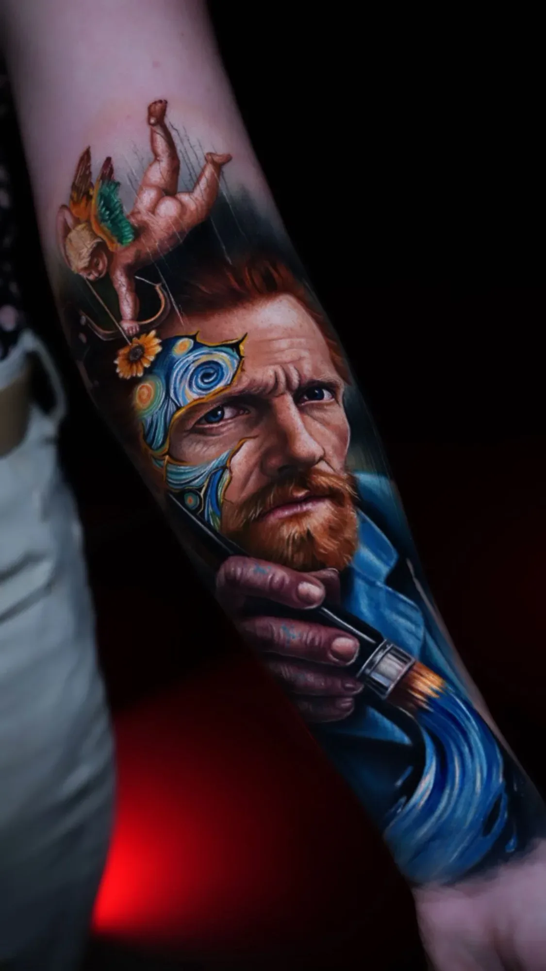 A hyperrealistic colour tattoo on a forearm showing a portrait of Vincent van Gogh with a paintbrush; swirling Starry Night patterns overlay the left side of his face, a yellow sunflower sits nearby, and a winged cherub with bow and arrow floats above.