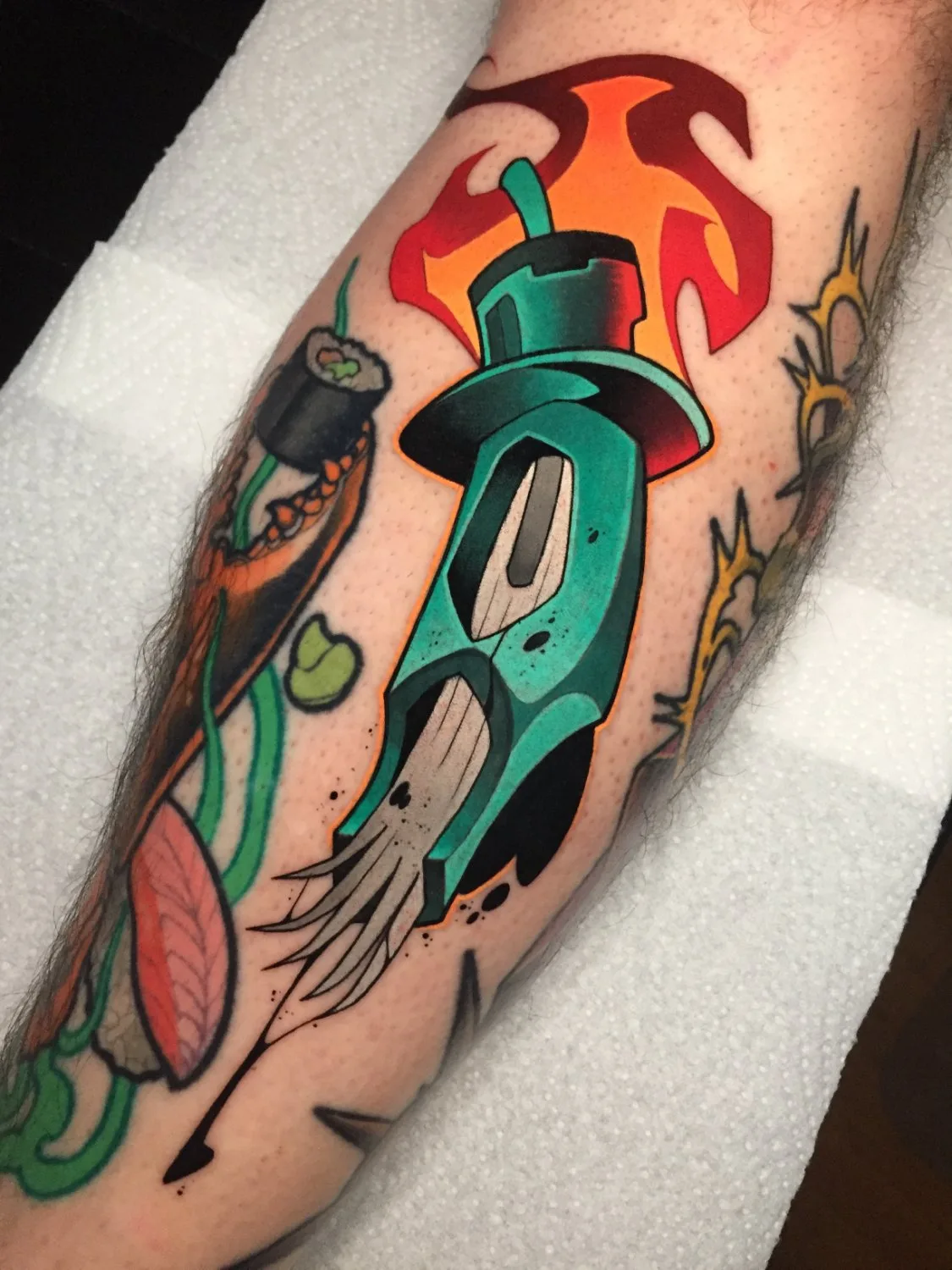 A forearm tattoo showing an anthropomorphic squid wearing a top hat with red flames above it, sushi rolls and seaweed to the left, and abstract orange and yellow spiked shapes to the right, all rendered in thick black outlines with vibrant turquoise, red, and green fills.