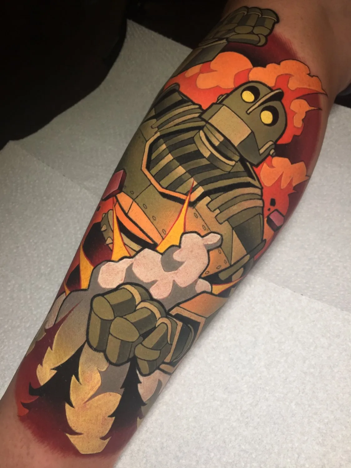 An arm tattoo depicting the Iron Giant from the chest up, rendered in olive green and muted brown with glowing yellow eyes, surrounded by dynamic flames in orange, red, and yellow, and soft white smoke, all executed in clean bold outlines with a comic-book illustrative quality.