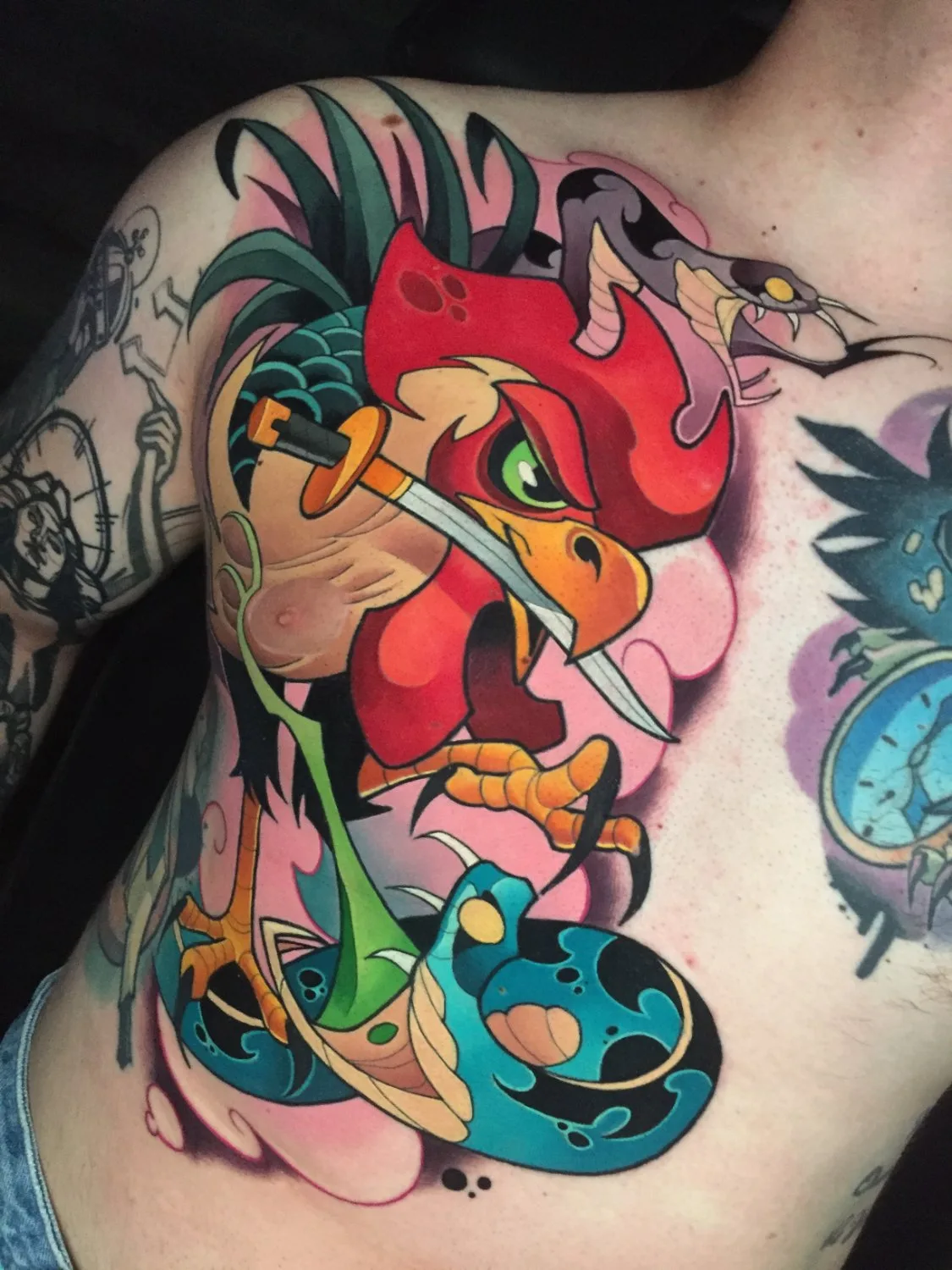 A chest tattoo showing a fierce rooster with green eyes and orange talons gripping a large blue-and-black coiled snake, a second grey snake arching over the rooster's comb, and a small silver sword piercing the bird's neck, rendered in bold outlines with rich reds, oranges, and greens on a pink back