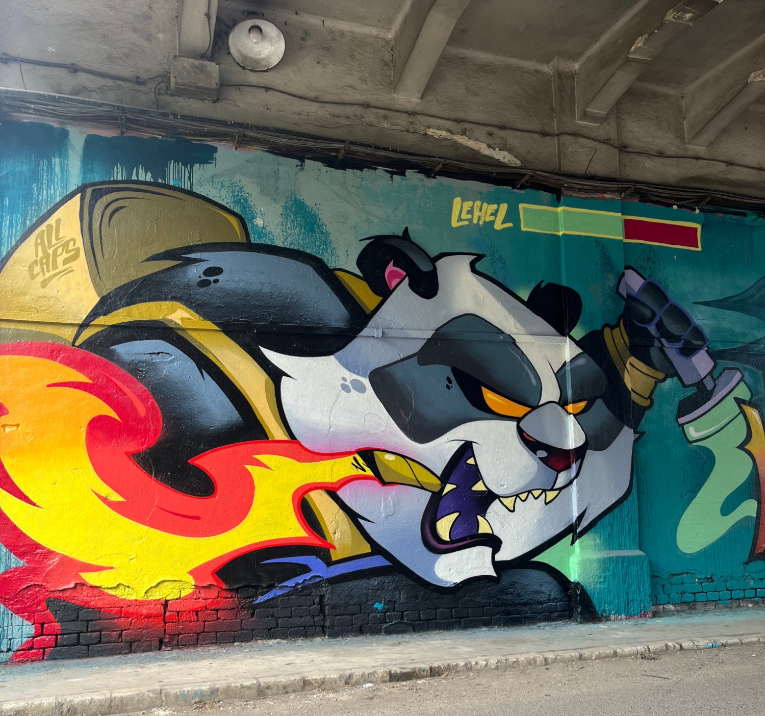 A large graffiti mural on a concrete wall under a bridge showing a black-and-white panda character in a yellow cape, exhaling red and yellow flames to the left and spraying green and yellow paint to the right, with 'LEHEL' written in yellow in the upper right.