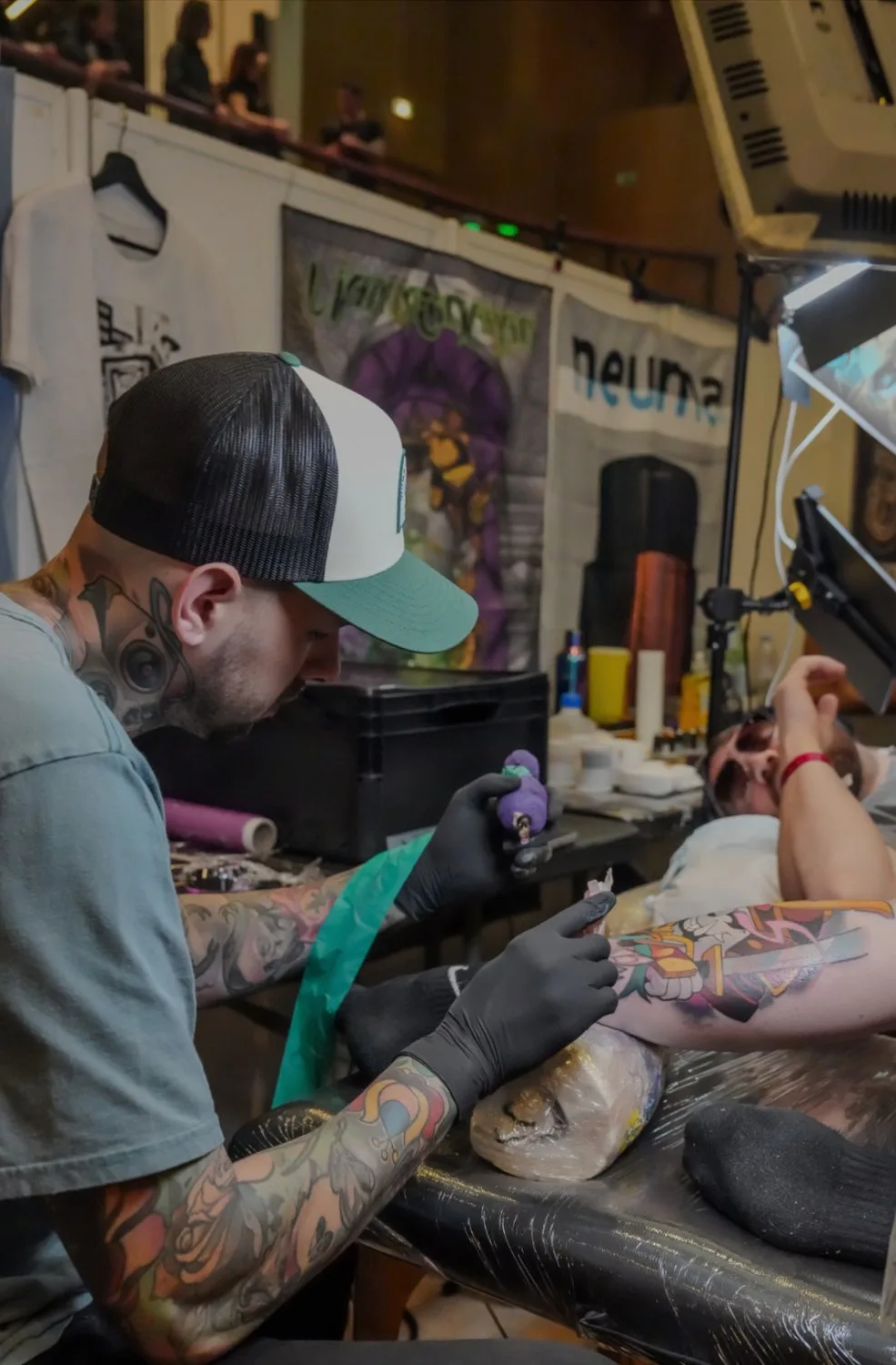 A tattoo artist wearing a teal t-shirt and black gloves leans over a client's leg at what appears to be a convention, applying a colourful cartoon-style tattoo; convention banners and ink bottles are visible in the blurred background.
