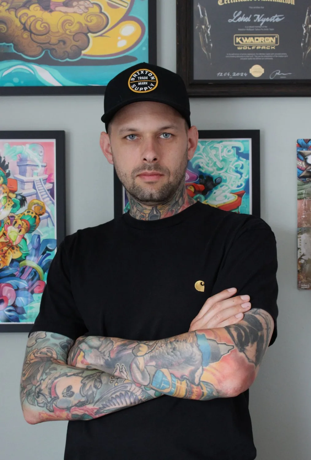 A tattooed man in a black cap and black t-shirt stands with arms crossed in front of a light grey wall hung with framed artworks and a certificate bearing the name Lehel Nyeste and the text KWADRON WOLFPACK with a date of 12.04.2024.