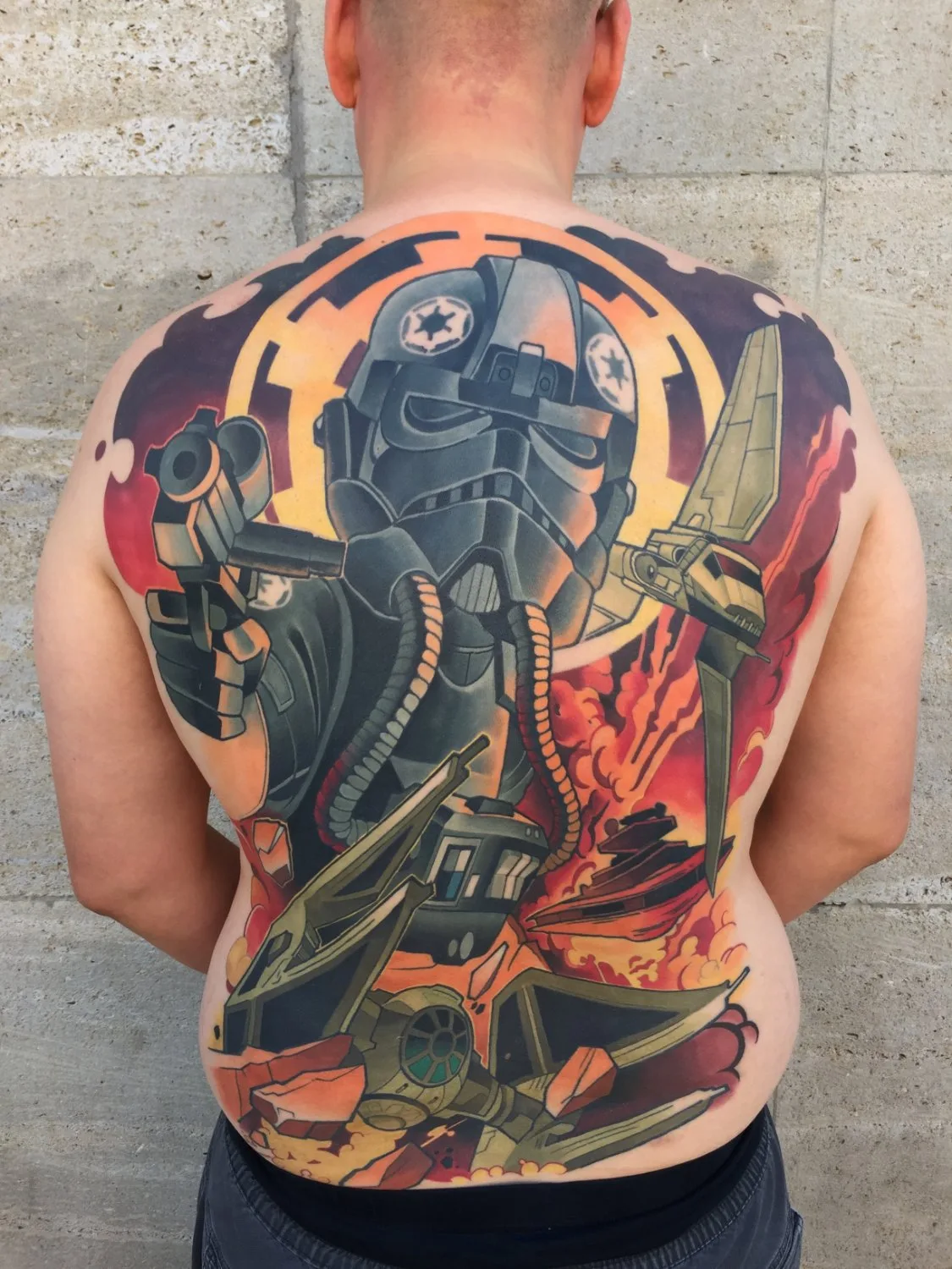A full back tattoo depicting a TIE fighter pilot from the chest up holding a blaster, flanked by several TIE fighters in various stages of flight and explosion, with intense orange, red, and yellow fire effects filling the background and dark grey and black tones defining the spacecraft and pilot's