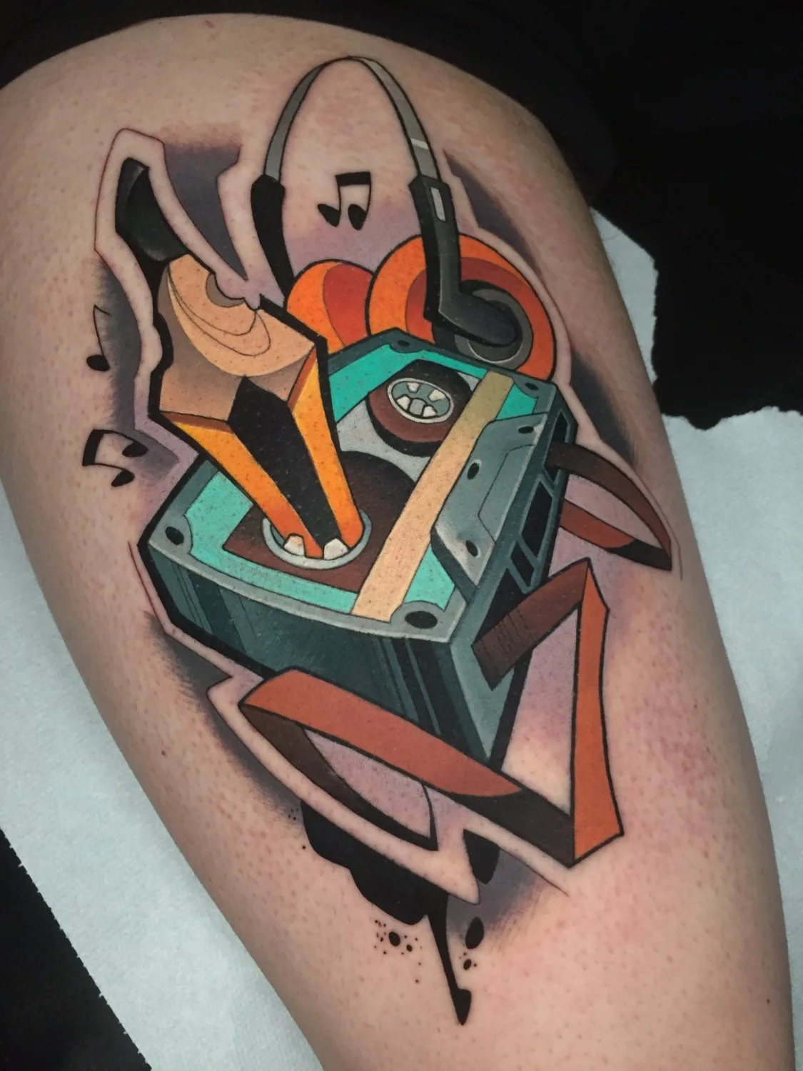 A tattoo on the leg showing a teal cassette tape at centre with a sharpened pencil emerging from its top, a pair of black headphones with orange earcups positioned above, musical notes scattered around, and abstract orange and black geometric shapes at the base, rendered in bold outlines with rich c