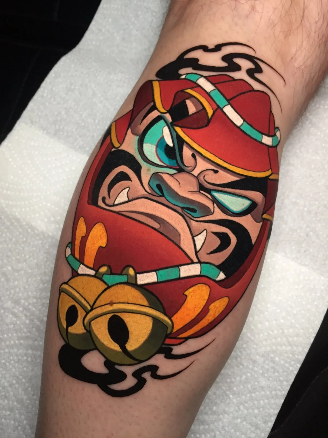 A leg tattoo depicting the face of a Daruma doll in deep red with teal eye and headband accents, two large golden bells hanging below the face, and black abstract flame or smoke shapes emanating from behind the bells.