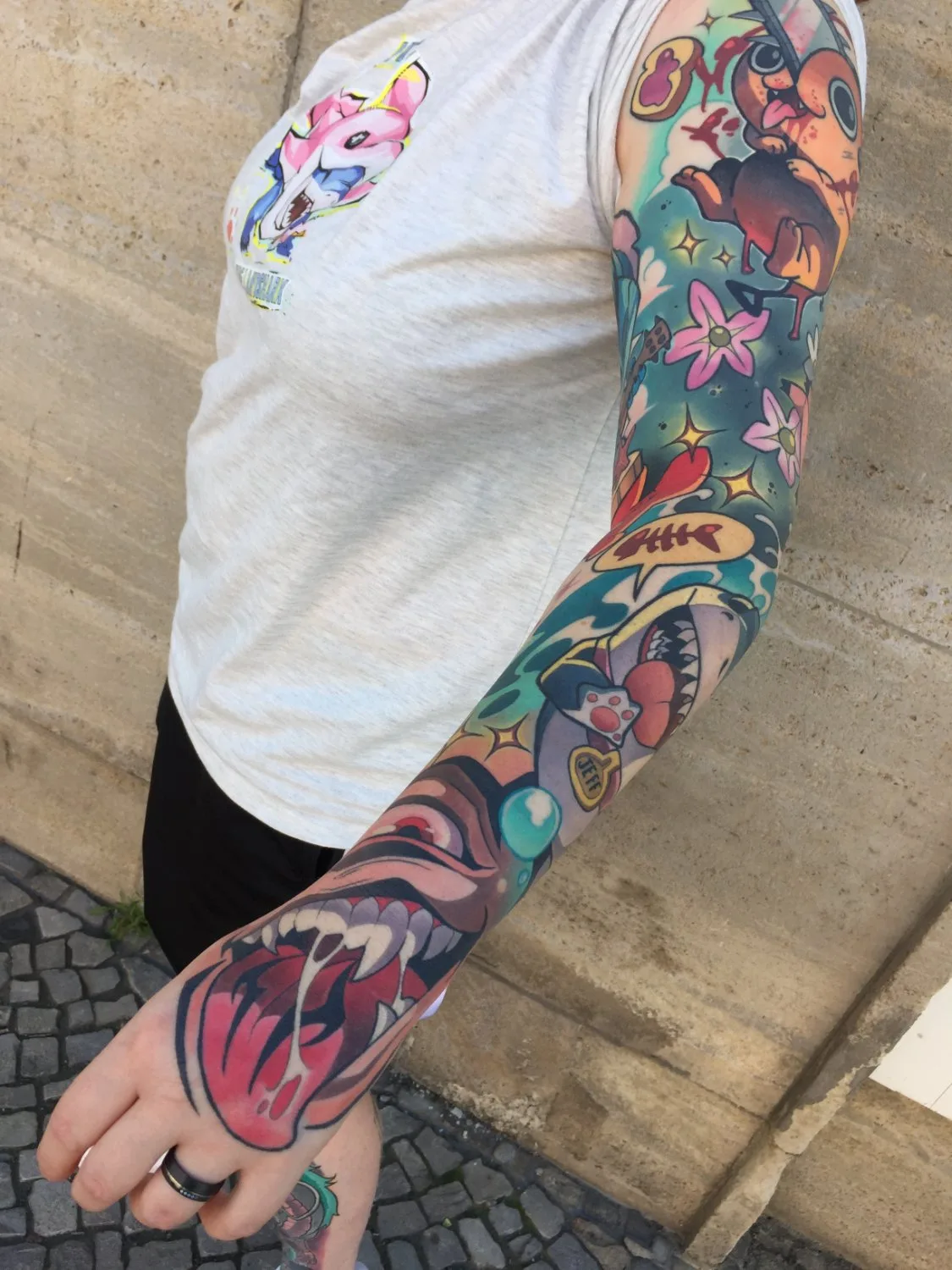 A full arm sleeve tattoo featuring a small reddish-brown creature with red splatters near the shoulder, pink stylised flowers on a teal background across the forearm, a fishbone in a speech bubble, a blue orb, and a large open mouth with sharp teeth and a dripping pink tongue near the wrist, all in