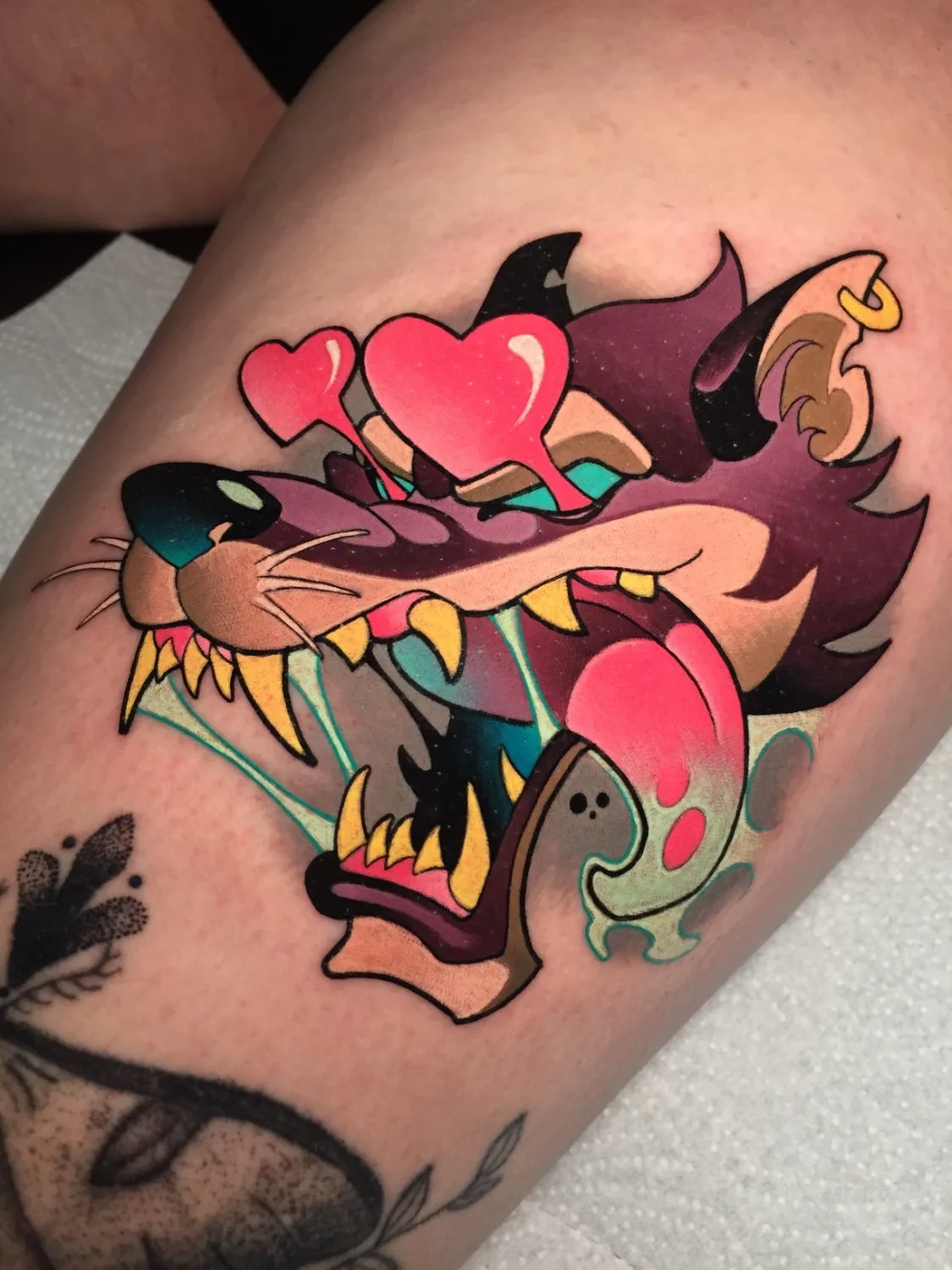 A tattoo depicting a wolf's head with an open snarling mouth, yellow teeth, and a bright pink tongue, wearing melting heart-shaped pink sunglasses, its fur shaded in deep purple and reddish-brown with lighter purple highlights, a small gold hoop earring visible, all outlined in bold black lines.