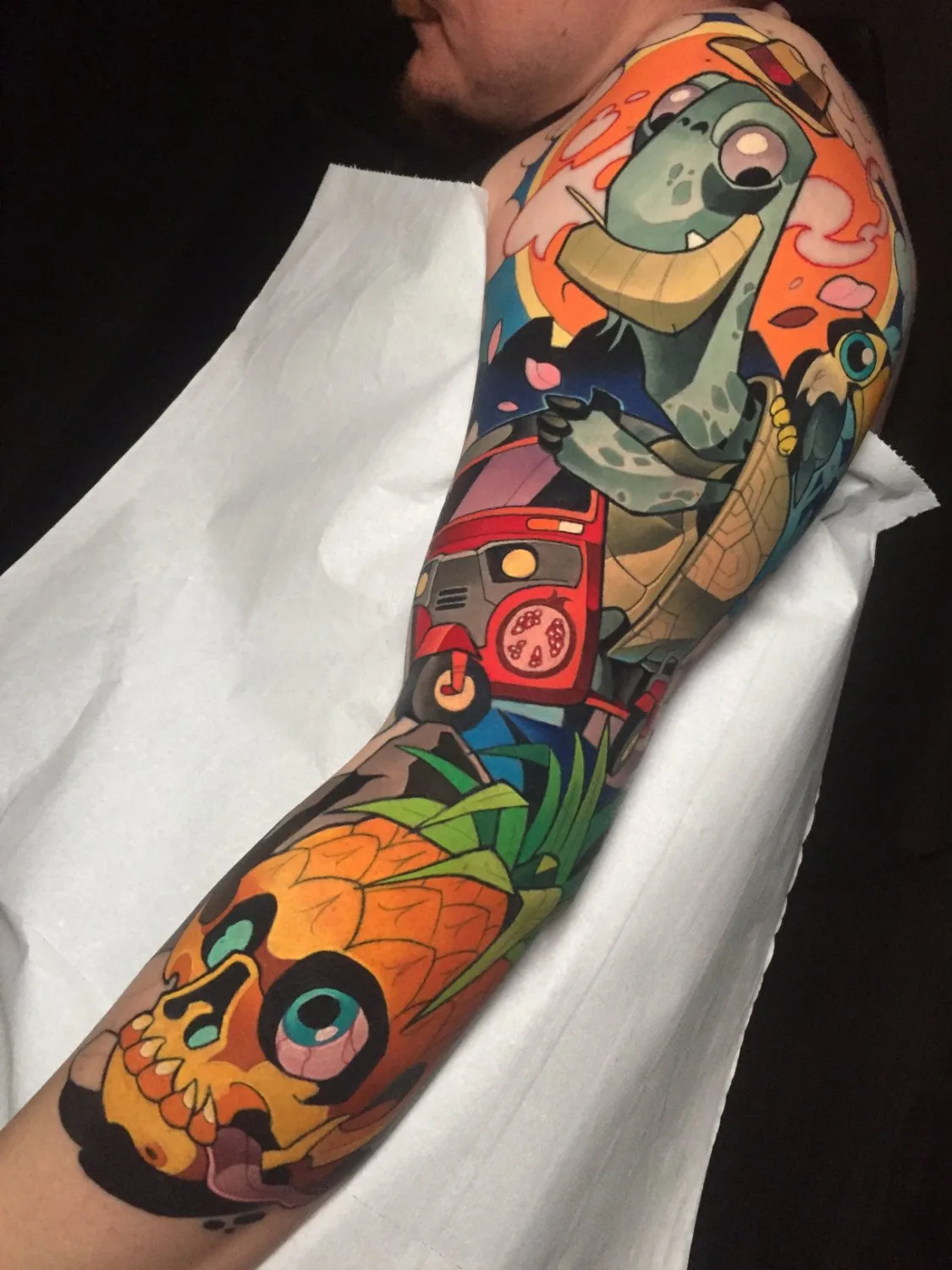 A full arm sleeve tattoo showing a light-green tortoise wearing a straw hat near the shoulder, a red three-wheeled vehicle below it, a small blue-eyed bird, and in the lower section a pineapple whose upper half becomes a skull with a blue eye and protruding tongue, all in bold cartoon outlines and v