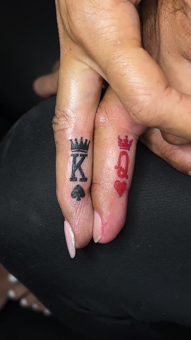 Close-up of two fingers held near each other: the left bears a bold black 'K' with a spade and crown, the right a red 'Q' with a heart and crown, referencing King and Queen playing card symbols.