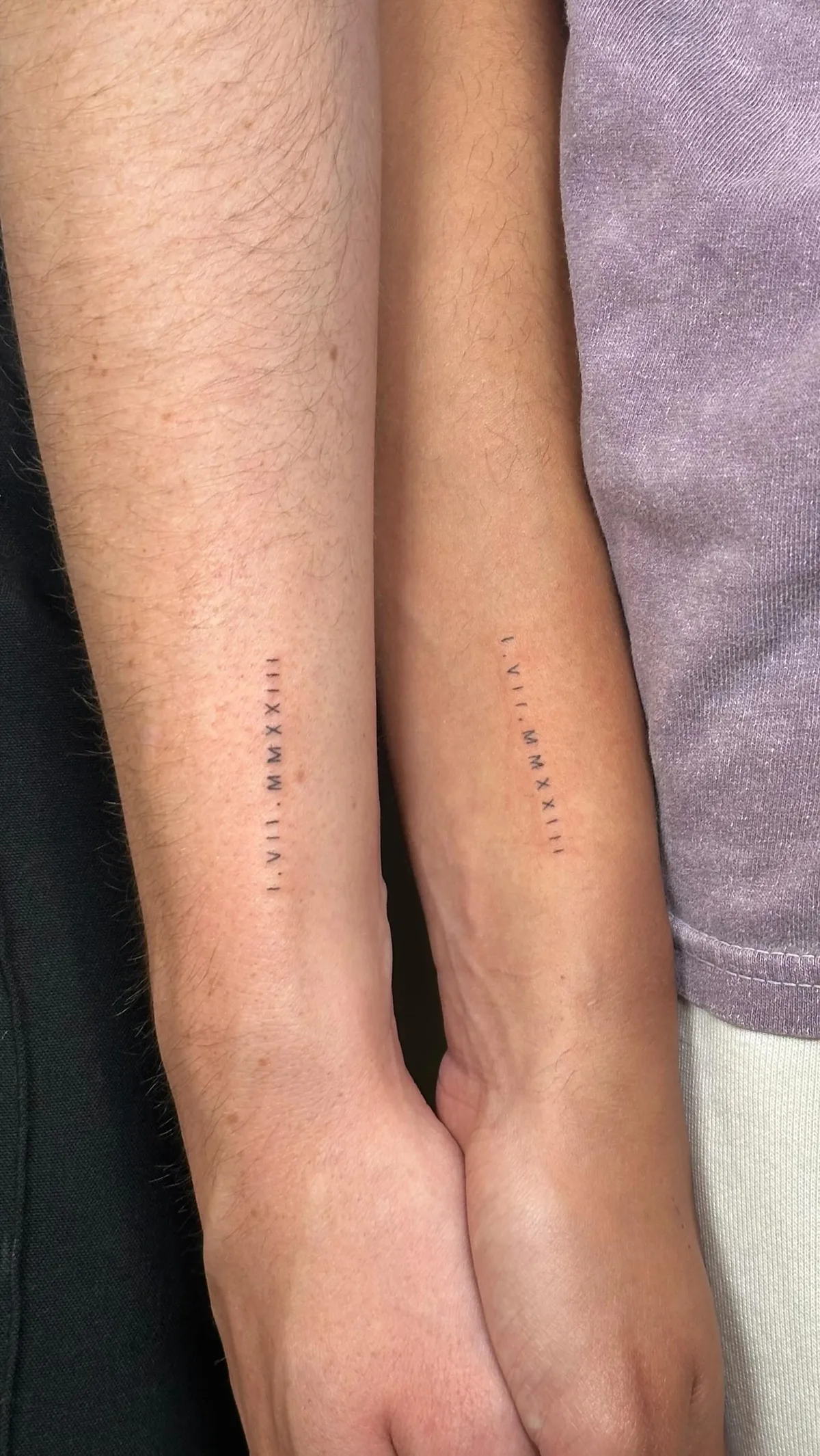 Two forearms placed side by side. The left bears the Roman numeral tattoo 'I. VII. MMXIII.' and the right bears 'VII. MMXIII.' Both are rendered in minimalist fine-line black ink, sharing the year 2013 as a common element.