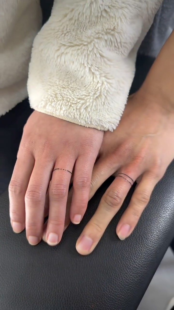 Two hands with matching minimalist thin black broken-circle ring tattoos on the ring fingers.