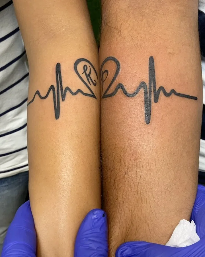 Two forearms placed side by side, each bearing a minimalist line-art tattoo where an ECG heartbeat line transitions into one half of a heart. When the arms meet, the two halves form a complete heart. The left half contains the initial 'R' in stylised script. Black ink on contrasting skin tones.