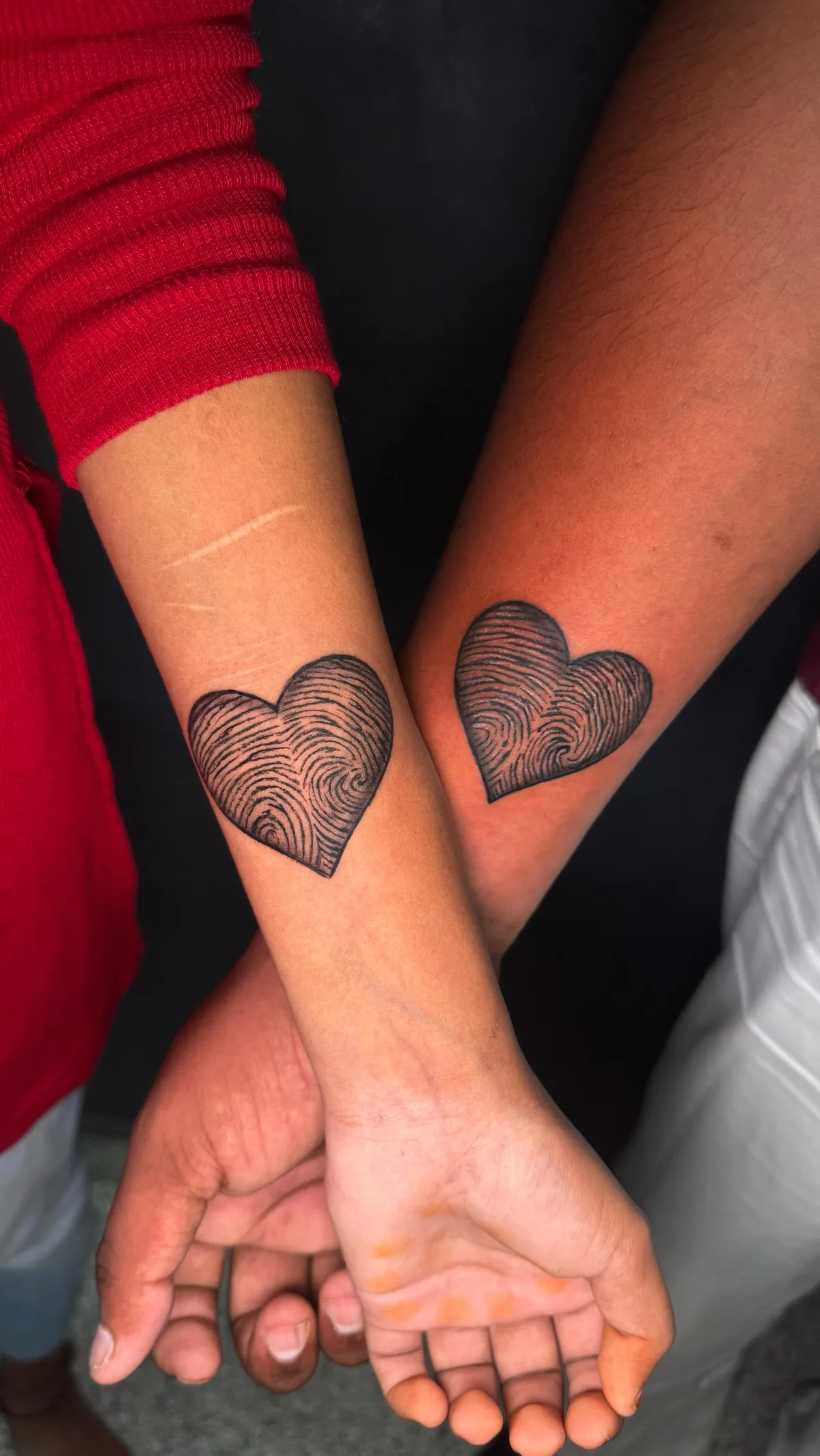 Two forearms held close together, each displaying a heart-shaped tattoo filled with intricate swirling lines that mimic fingerprint ridges and whorls. Rendered in fine-line blackwork against a deep black background. The hands are partially visible at the bottom, fingers intertwined.
