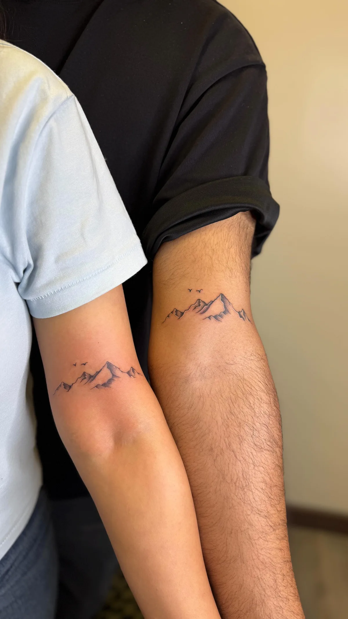 Two forearms angled toward each other, each bearing a fine-line black tattoo of a mountain range with small V-shaped bird silhouettes in flight above the peaks. Subtle shading gives dimension to the mountains.
