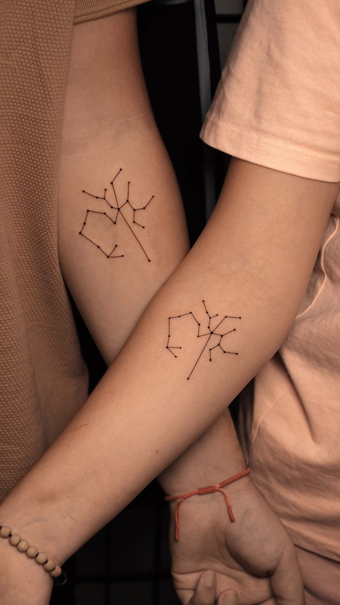 Two forearms placed close together, each bearing a minimalist constellation tattoo made of small black dots connected by thin straight lines. The two constellations are distinct but share the same visual language. One wearer has a wooden bead bracelet; the other has a red string bracelet.