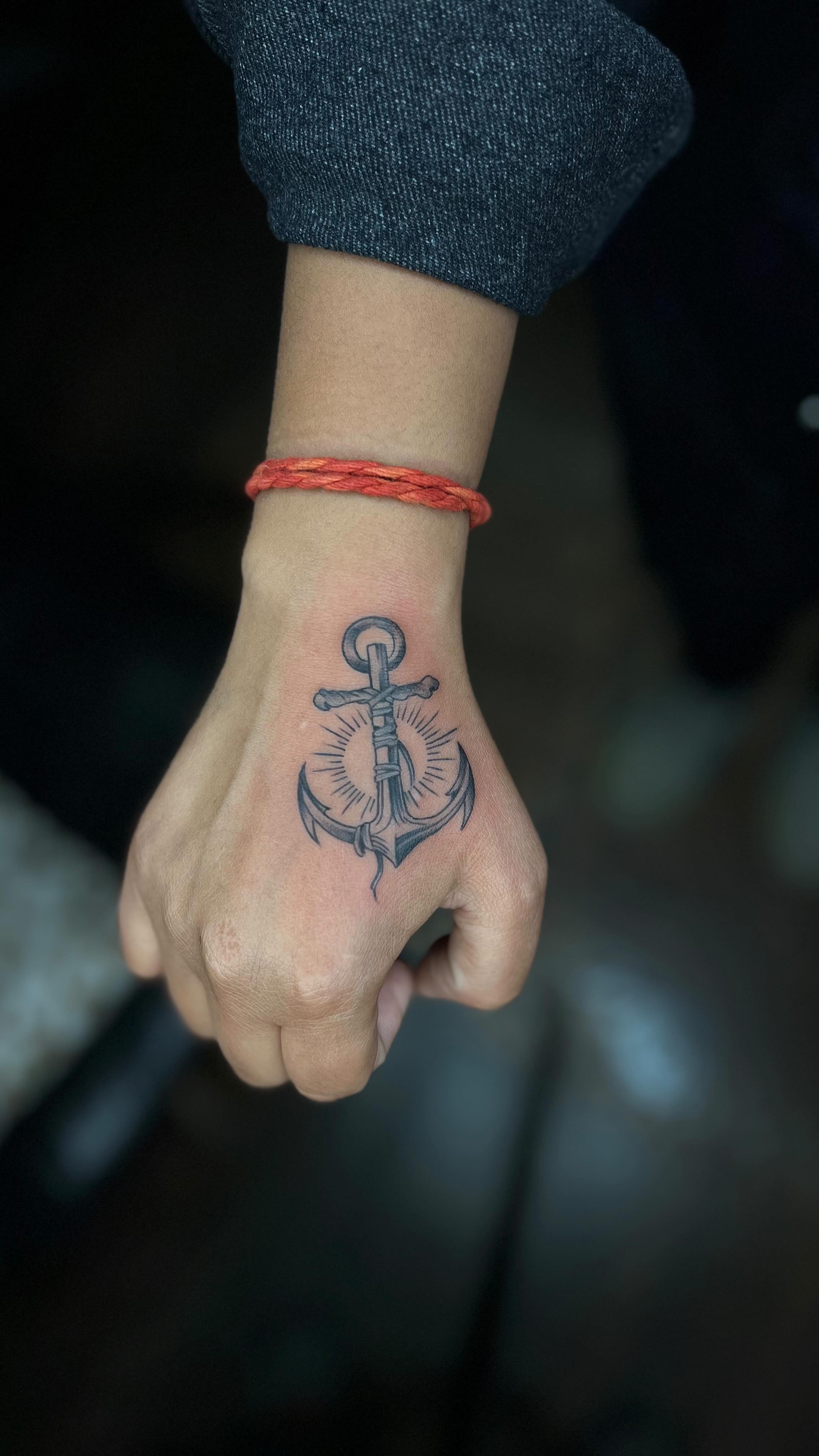 A black and grey tattoo of a traditional anchor with radiating lines behind it, placed on the fleshy area of the hand between the thumb and index finger. A braided red cord bracelet is visible on the wrist above.