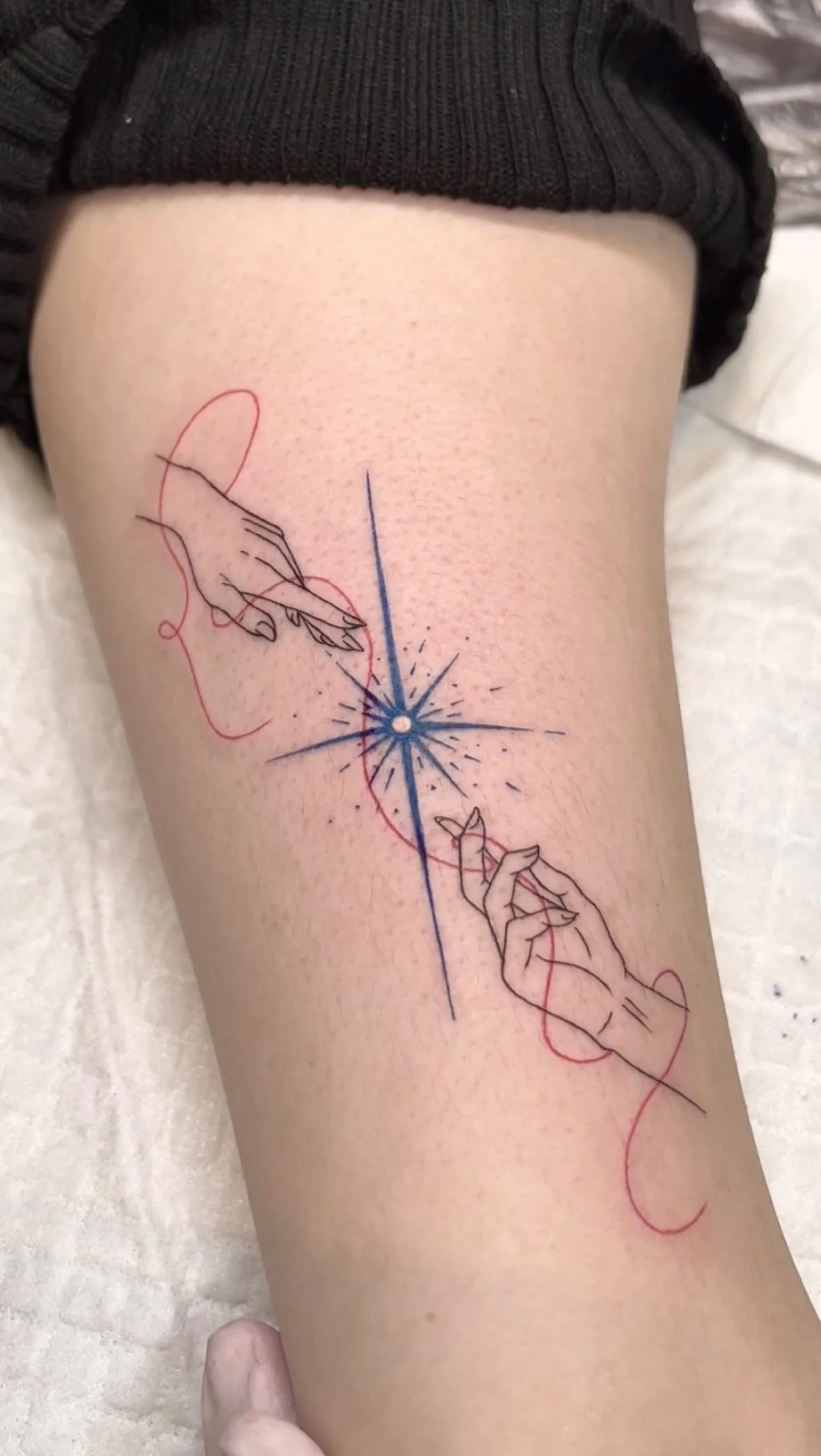 A forearm tattoo showing two minimalist outlined hands reaching toward a stylised blue compass star. A bright red thread connects the fingers and the star's centre, referencing the red thread of fate. Thin black linework with limited colour.
