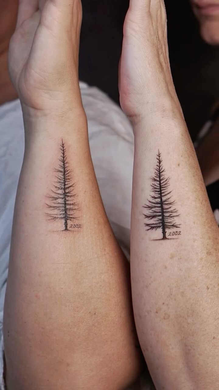 Two forearms side by side, each bearing a fine-line minimalist tattoo of a bare coniferous tree standing on a small patch of ground. Below each tree the year '2002' is inscribed in small sans-serif lettering. Black ink on lightly freckled skin.