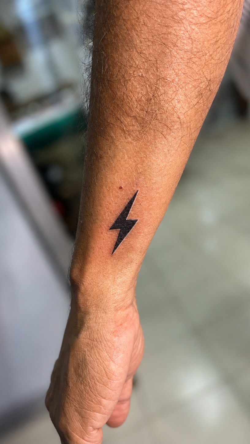 A minimalist solid black lightning bolt tattoo on the inner forearm near the wrist. The design is a clean, sharp silhouette with no shading or additional detail.