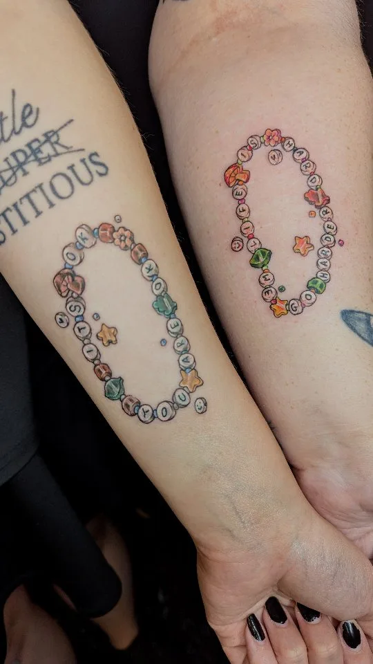Two forearms placed side by side, each bearing a neo-traditional tattoo of a beaded friendship bracelet. The left bracelet spells 'I LOVE YOU OKAY' and the right spells 'I LOVE YOU HARDER', both interspersed with charm beads depicting stars, flowers, mushrooms and bells. Bold black outlines with vib
