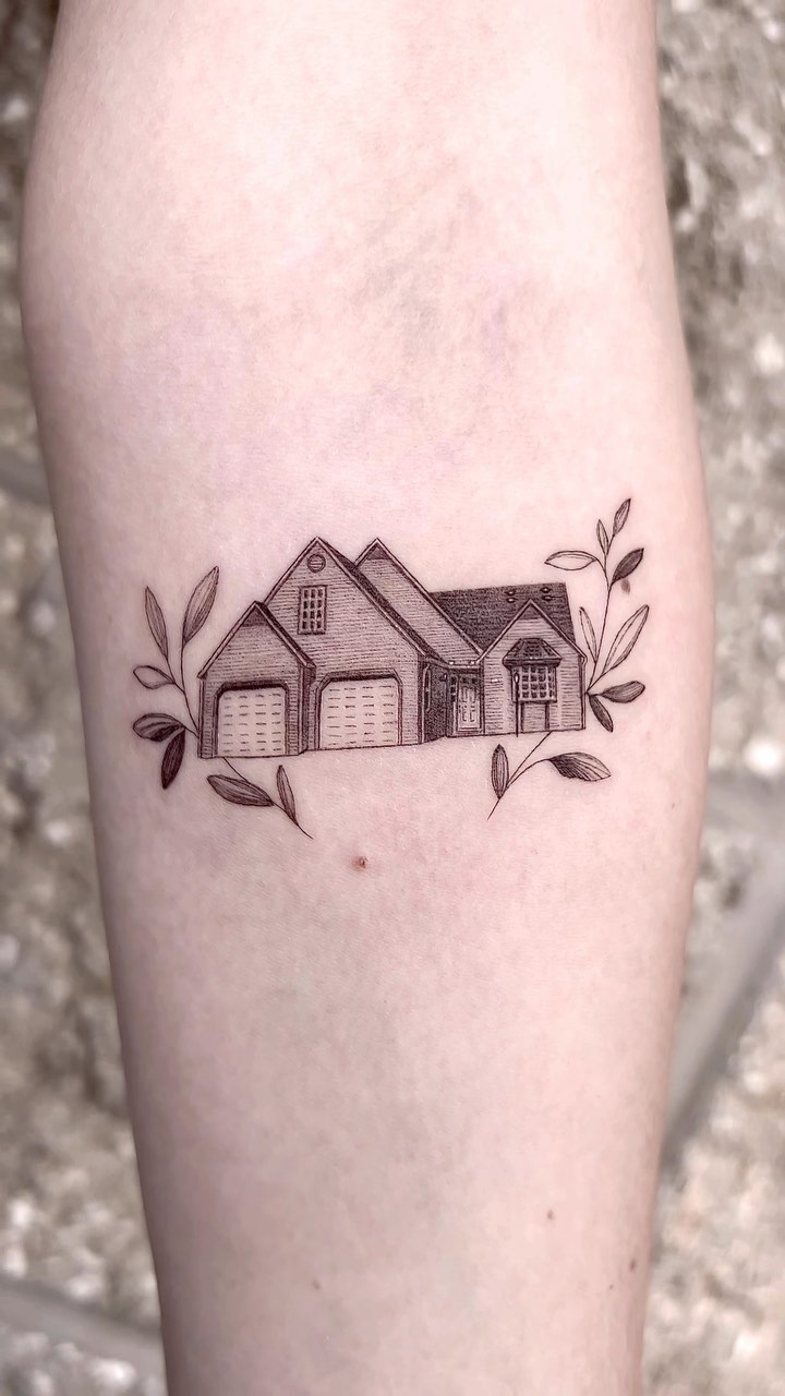 A fine-line black and grey tattoo of a detailed suburban house with a gabled roof, windows and garage doors, flanked symmetrically by delicate leaf sprigs. Rendered in micro-realism style with subtle hatching and stippling for depth.
