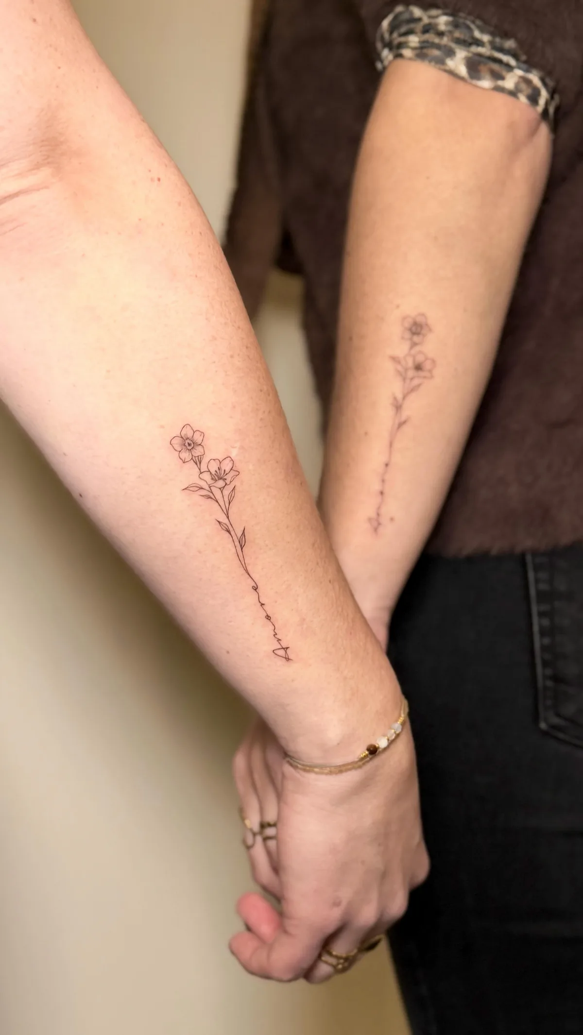 Two forearms held together with hands clasped. Each forearm bears a fine-line tattoo of a winding stem with delicate flowers and leaves. Below each floral design, the word 'Amore' is written in flowing cursive script. Black ink on fair skin.