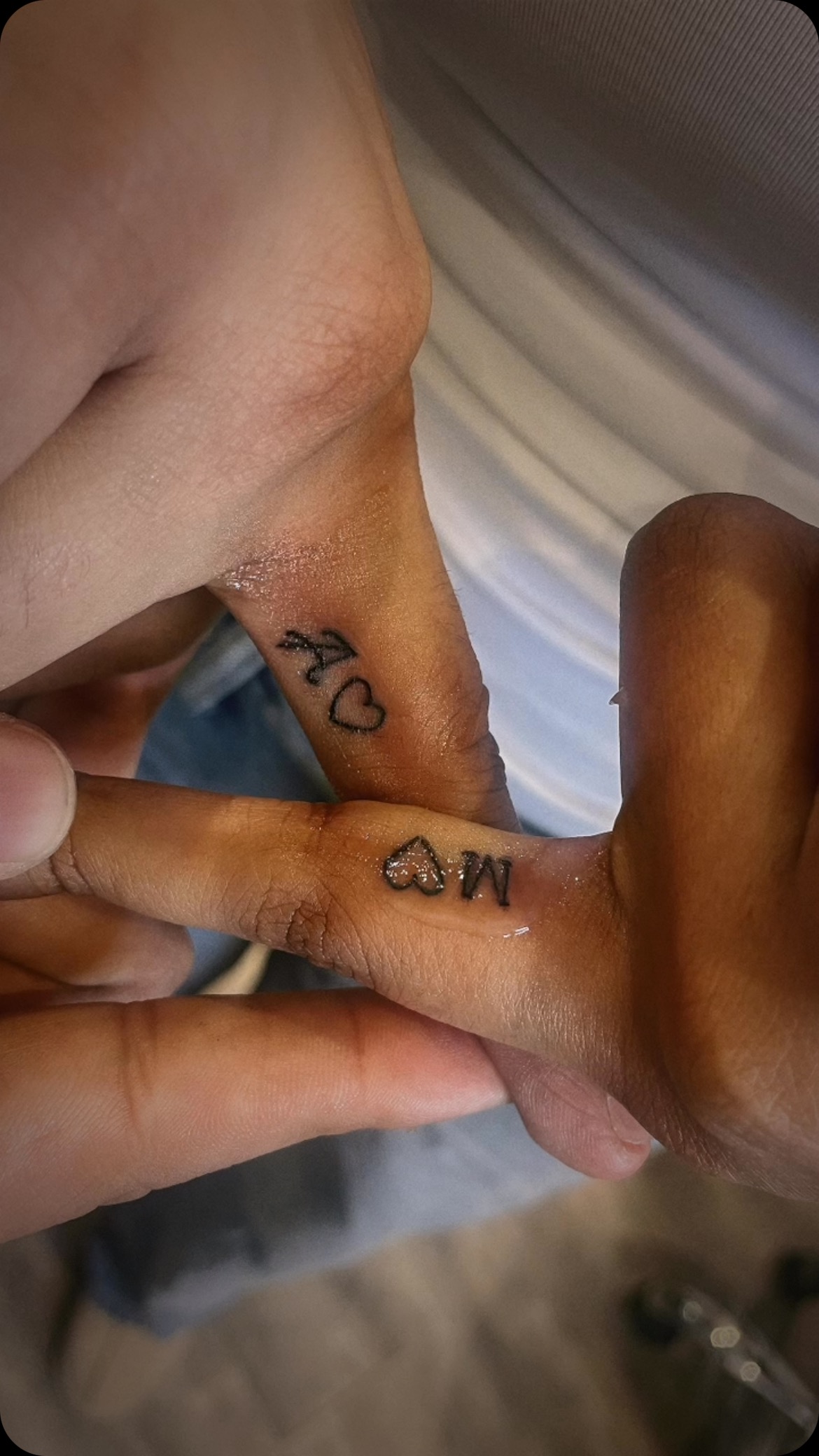 Two fingers of different people, each with a letter and a small heart tattooed on the side