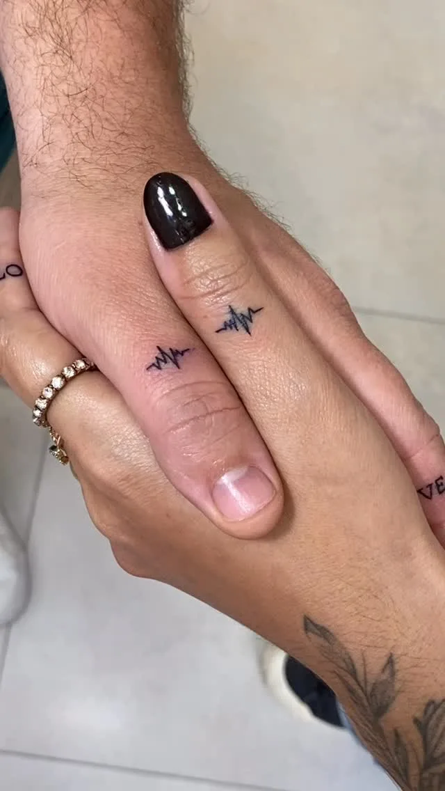 Two individuals' hands intertwined showing fine-line minimalist tattoos on fingers and forearms, with black nail polish visible on one thumb.