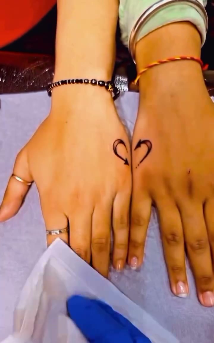 Two hands each bearing a small matching tattoo of a heart with an arrow through it, positioned near the base of the index and middle fingers.