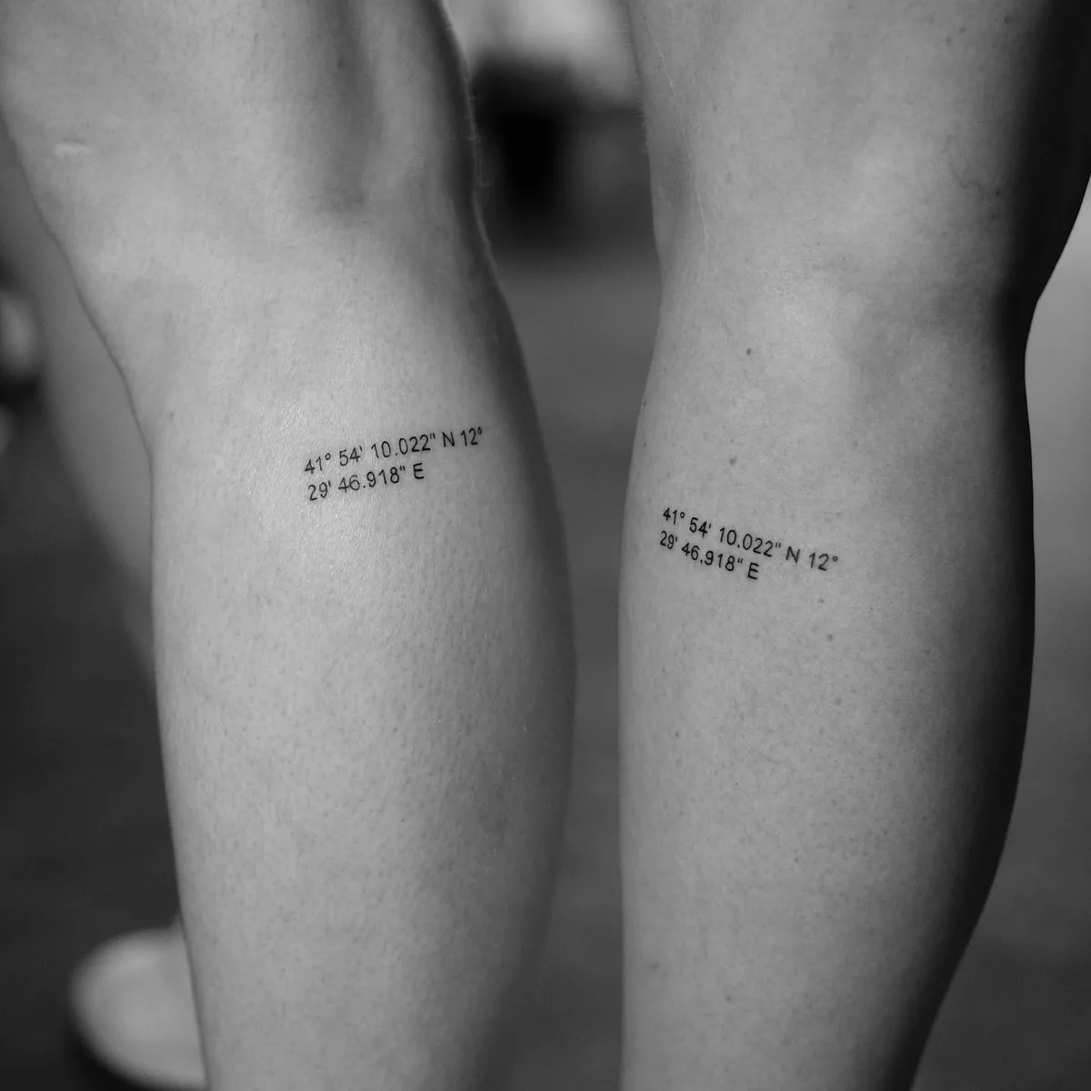 Black and white close-up of two lower legs, each bearing an identical small sans-serif tattoo of geographic coordinates reading '41° 54' 10.022" N 12° 29' 46.918" E'. The tattoos are placed symmetrically on the inner calves.