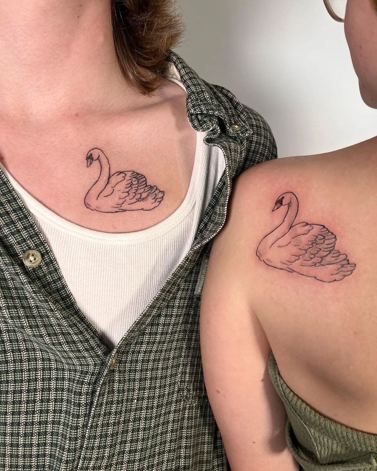 Two individuals shown from the chest and shoulder. One has a minimalist black line-art swan tattoo on the upper chest below the collarbone; the other has the same design on the upper back near the shoulder. Both swans face left with necks gracefully curved.