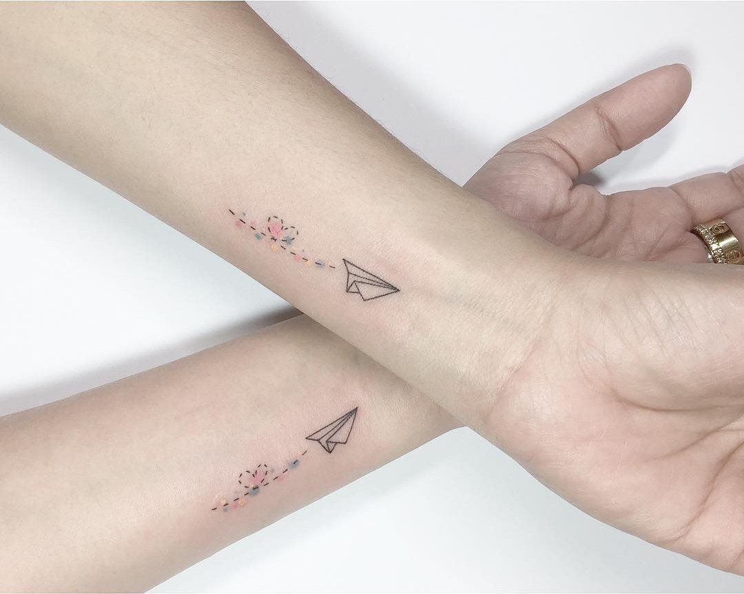 Two inner wrists each bearing a fine-line tattoo of a folded paper airplane trailing a dotted line interspersed with small pastel-coloured dots in pink, yellow and blue, with a loose flower-like cluster at the start of the trail. Black linework with soft colour accents.