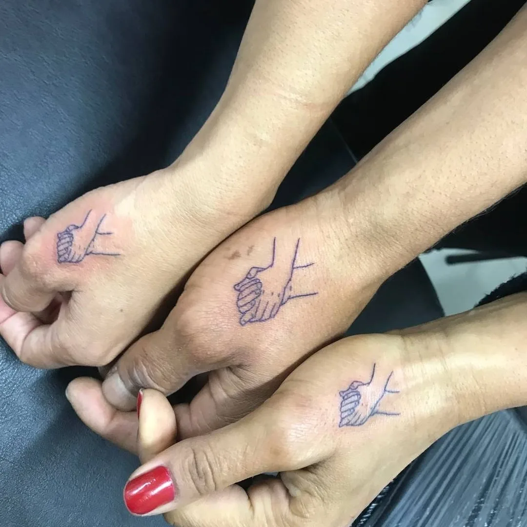 Three hands clasped together, each bearing a minimalist thin dark-blue line-drawing tattoo of hands holding each other. One fingernail is painted vivid red. The background is a dark textured surface.