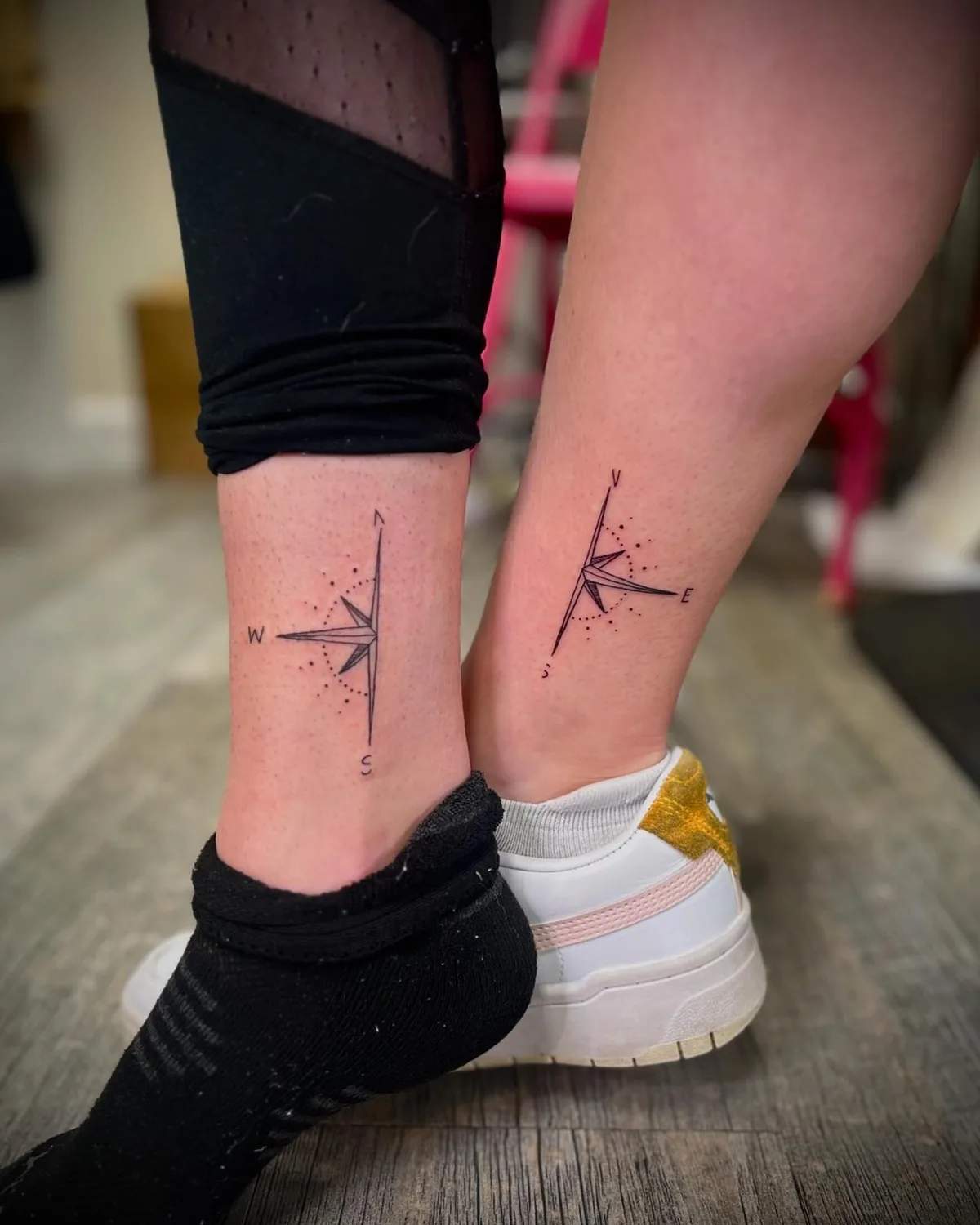 Close-up of two lower legs and ankles, each bearing a small fine-line black compass rose tattoo with cardinal direction letters and scattered dot accents. One leg wears black leggings; the other has a white and gold sneaker visible.
