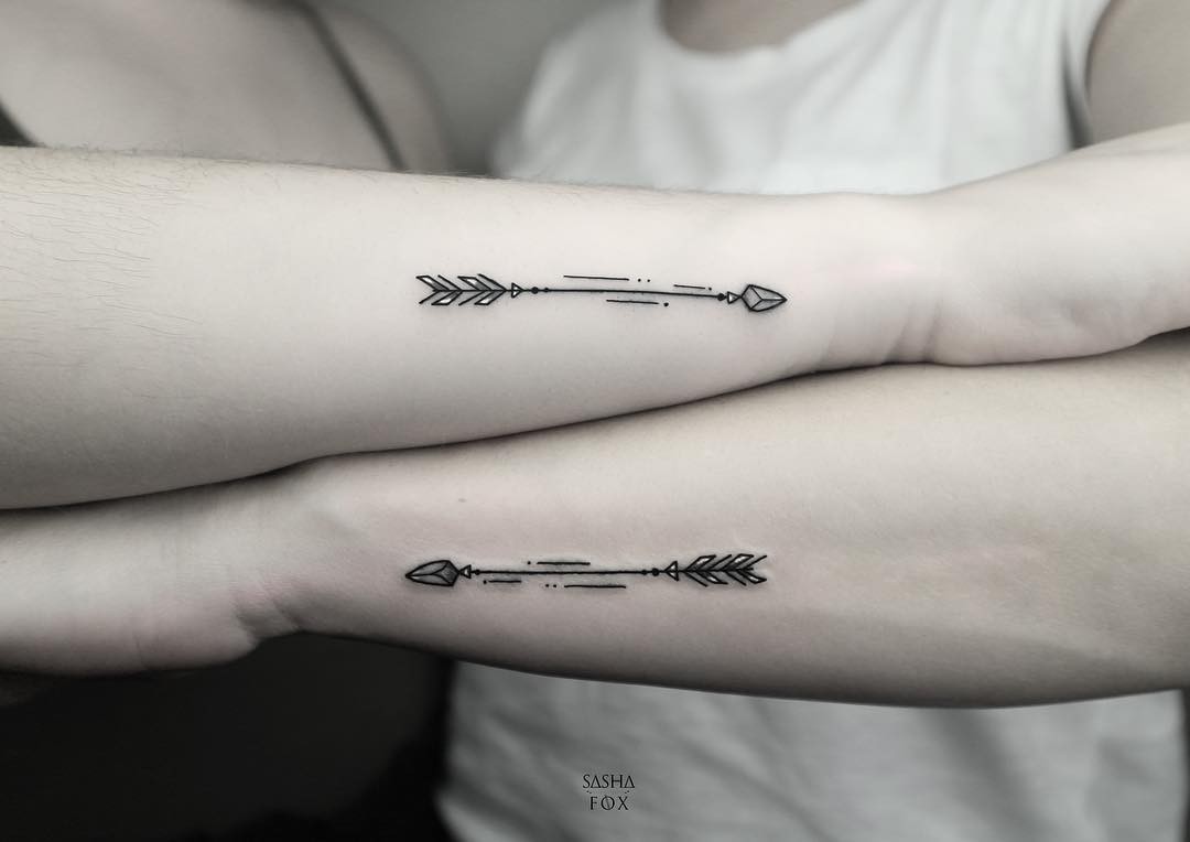 Two forearms crossed over each other, each bearing a minimalist geometric arrow tattoo in black ink. The arrows mirror each other in orientation, each featuring a chevron fletching, a thin shaft with dots and dashes, and a faceted arrowhead.