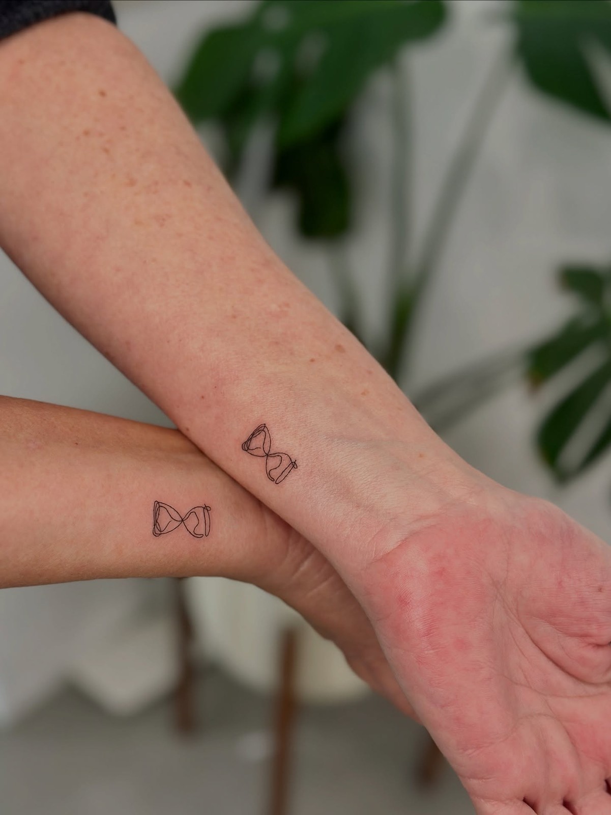 Two wrists held close together, each bearing a minimalist fine-line hourglass tattoo drawn in a continuous single-line technique. Black ink on fair skin with some freckles visible. A soft green plant is blurred in the background.