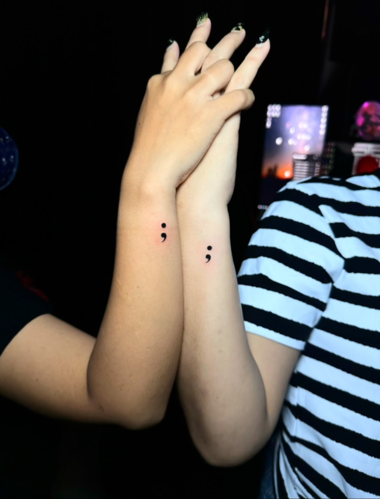Two forearms with hands clasped, each displaying a small black semicolon tattoo on the inner wrist. The tattoos are placed symmetrically and rendered in clean black ink on light skin.