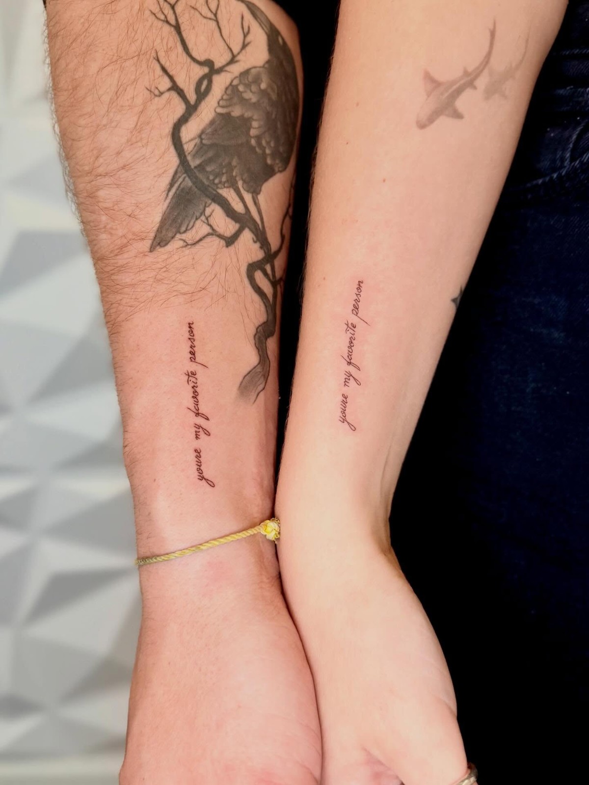 Two forearms held together, each bearing the phrase 'you're my favorite person' written vertically in delicate cursive script. One arm also features a detailed black and grey bird-on-branch tattoo above the text; the other has faint minimalist shark silhouettes higher up.