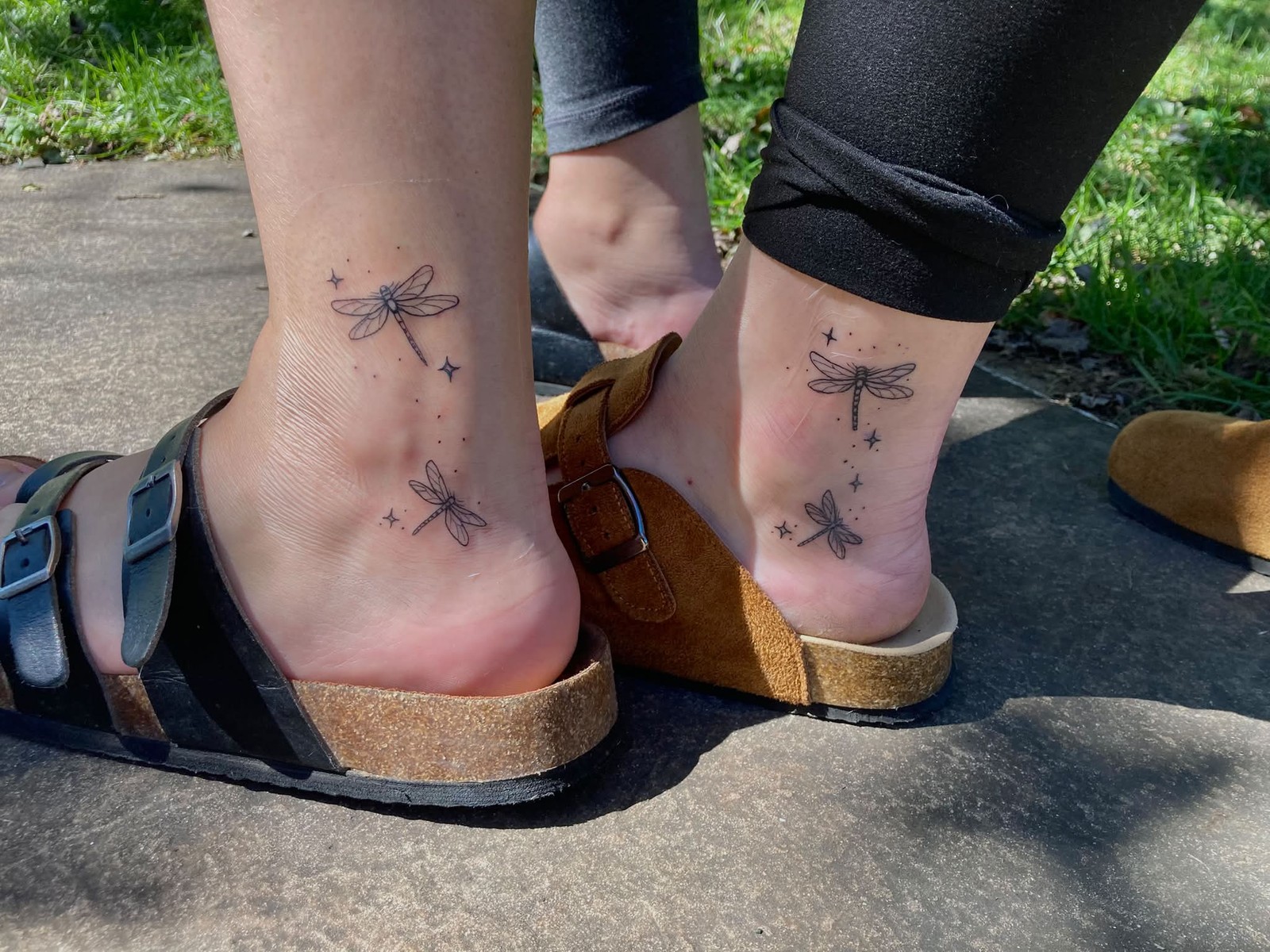Two ankles placed close together, each bearing a fine-line black tattoo of two dragonflies arranged vertically with small four-pointed stars scattered around them. The designs are delicate and minimal.