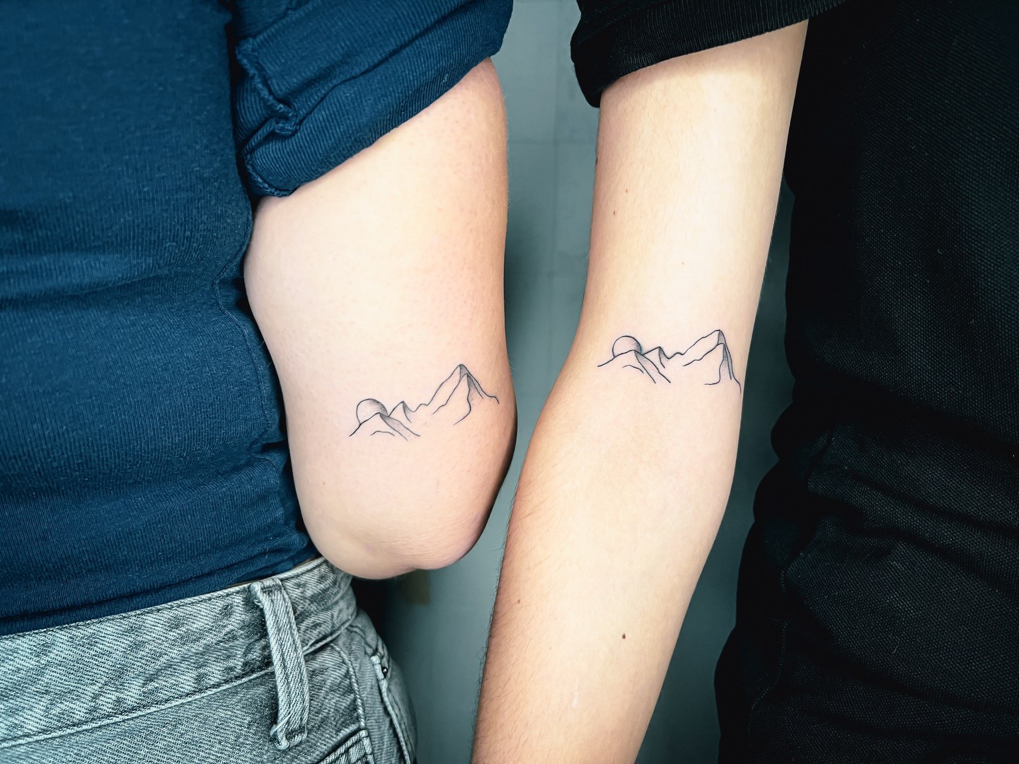 Two inner forearms placed side by side, each bearing a minimalist fine-line tattoo of jagged mountain peaks with a partial circle representing a rising sun behind them. Single-stroke black lines on lightly tanned skin.