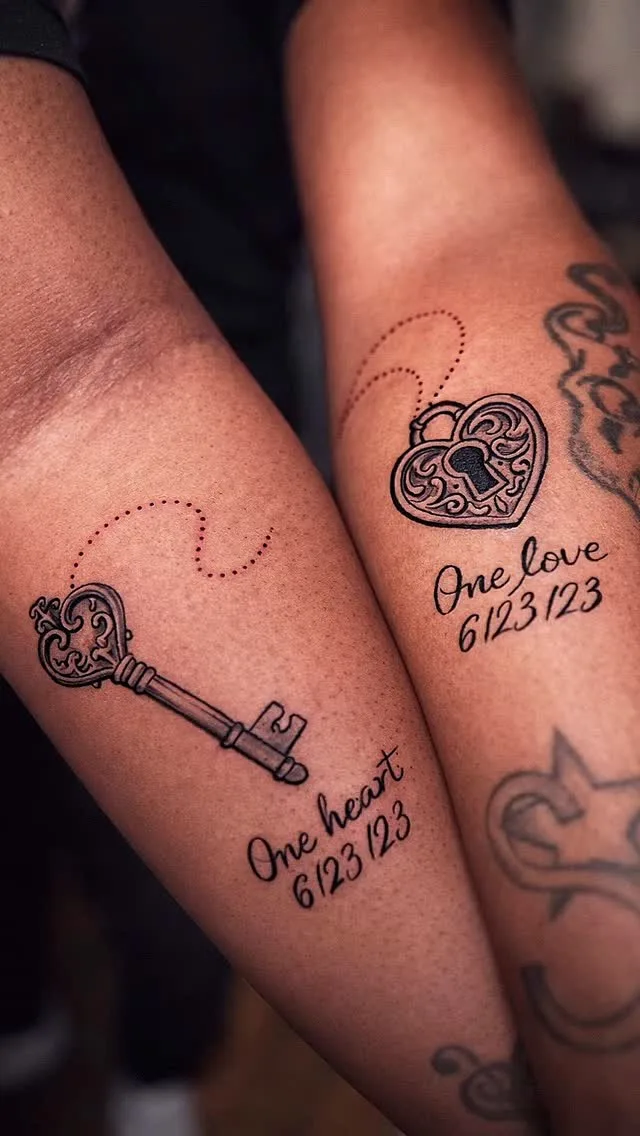 Two forearms angled toward each other. The left bears an ornate fine-line key tattoo with the text 'One heart' and date '6/23/23' beneath it. The right bears a heart-shaped locket with a keyhole and the text 'One love' and the same date. A dotted line visually connects the two designs.