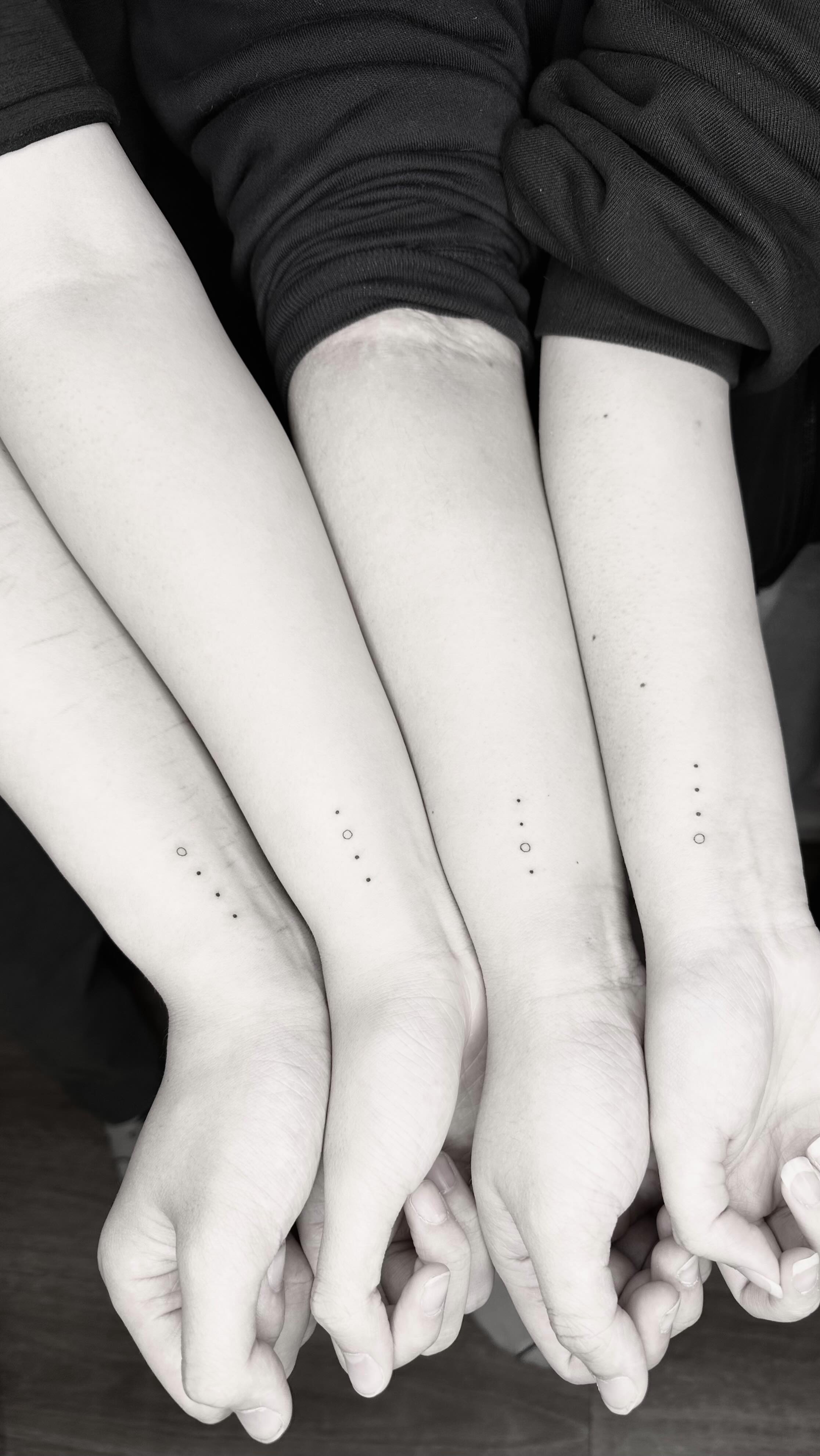 Black and white close-up of four forearms held together. Each forearm bears a unique small dotwork constellation pattern made of black dots and a single open circle. The hands are linked at the bottom of the frame, suggesting a family or group bond.