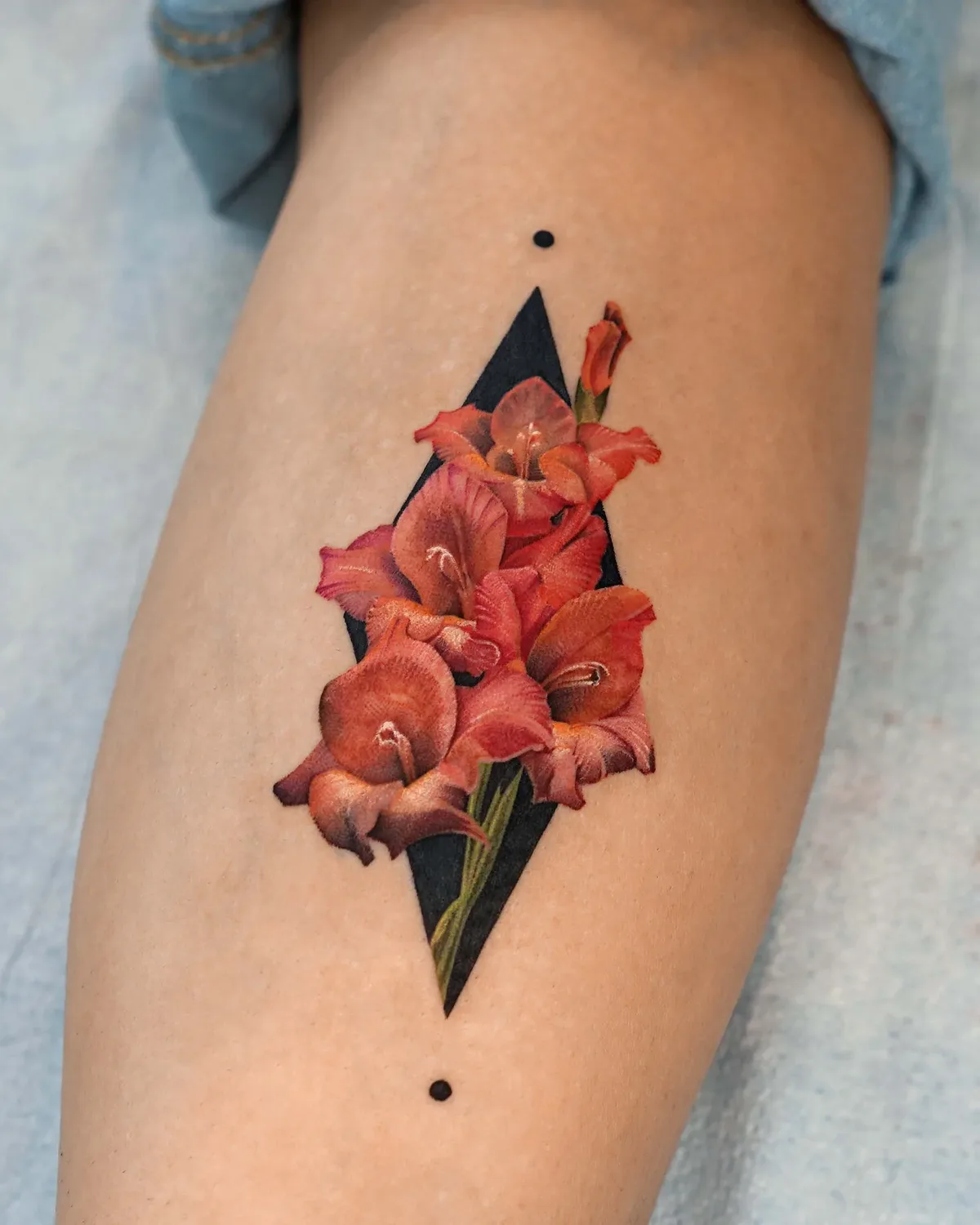 A tattoo on a leg showing four to five gladiolus blooms in shades of red, orange, and pink arranged inside a vertically oriented black diamond shape, with two small black dots above and below the diamond's points.