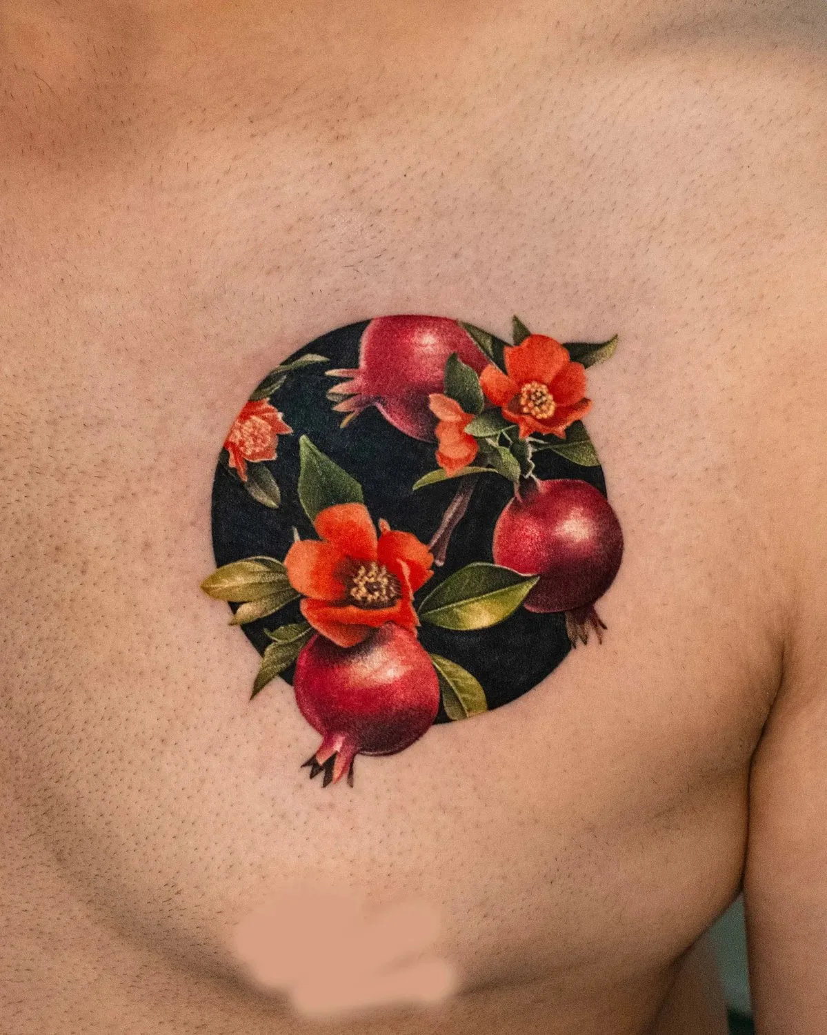 A close-up of a chest tattoo showing three ripe pomegranates with crown-like calyxes alongside vivid orange-red ruffled flowers and deep-green leaves, all arranged within a circular dark background.